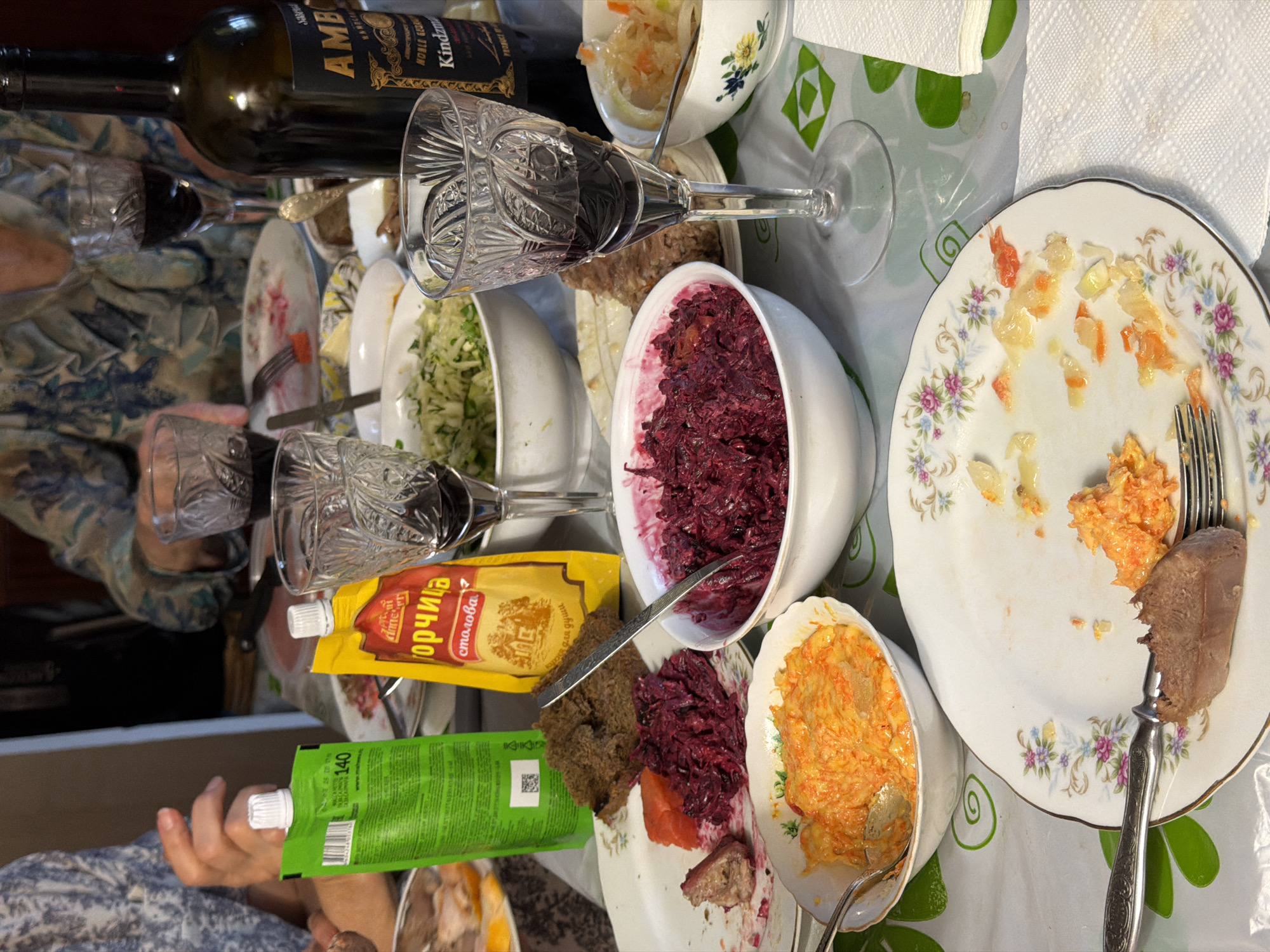 The image depicts a table laden with food, possibly in Krasnoyarsk, Russia, given the presence of Russian-branded products. The table is covered with a patterned tablecloth. Dishes containing various salads (beetroot, cabbage, carrot), cold cuts, and bread are scattered around. There are also tubes of mustard and horseradish, a bottle of wine, and crystal glasses filled with a dark liquid, presumably wine. A plate with food remnants and a fork suggests someone has been eating. Parts of two people are visible on either side of the table. The overall impression is of a feast or a casual meal in a home setting.
