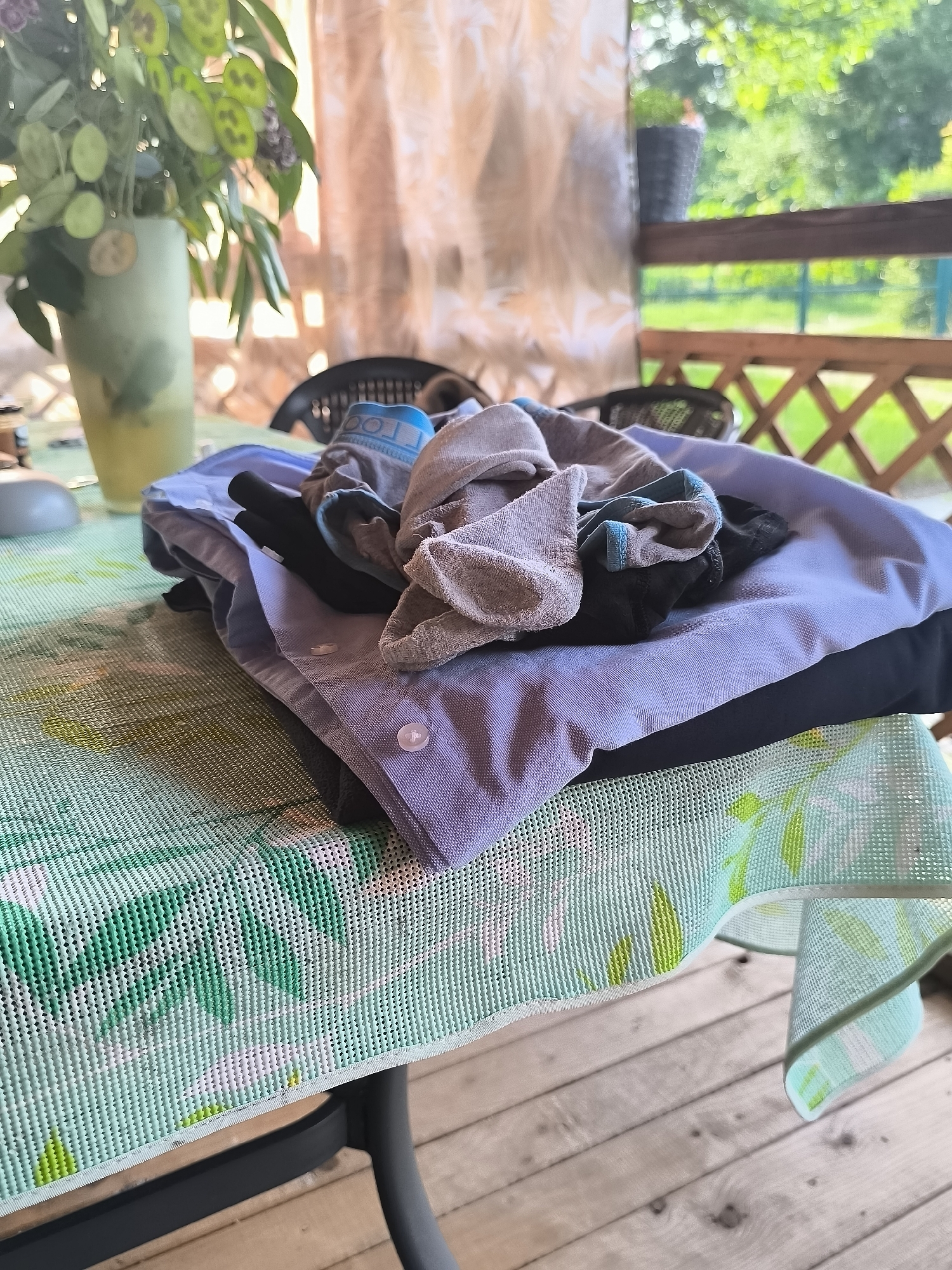A pile of folded clothes rests on a table covered with a green and white patterned tablecloth. The pile includes a light blue collared shirt, black fabric, and some gray socks. The table is situated on a wooden deck, with a blurred background of greenery and a fence. A vase with green plants is visible to the left of the pile. The scene appears to be outdoors, possibly on a porch or patio. No people are present. The location is identified as Dercekliai, Lithuania.