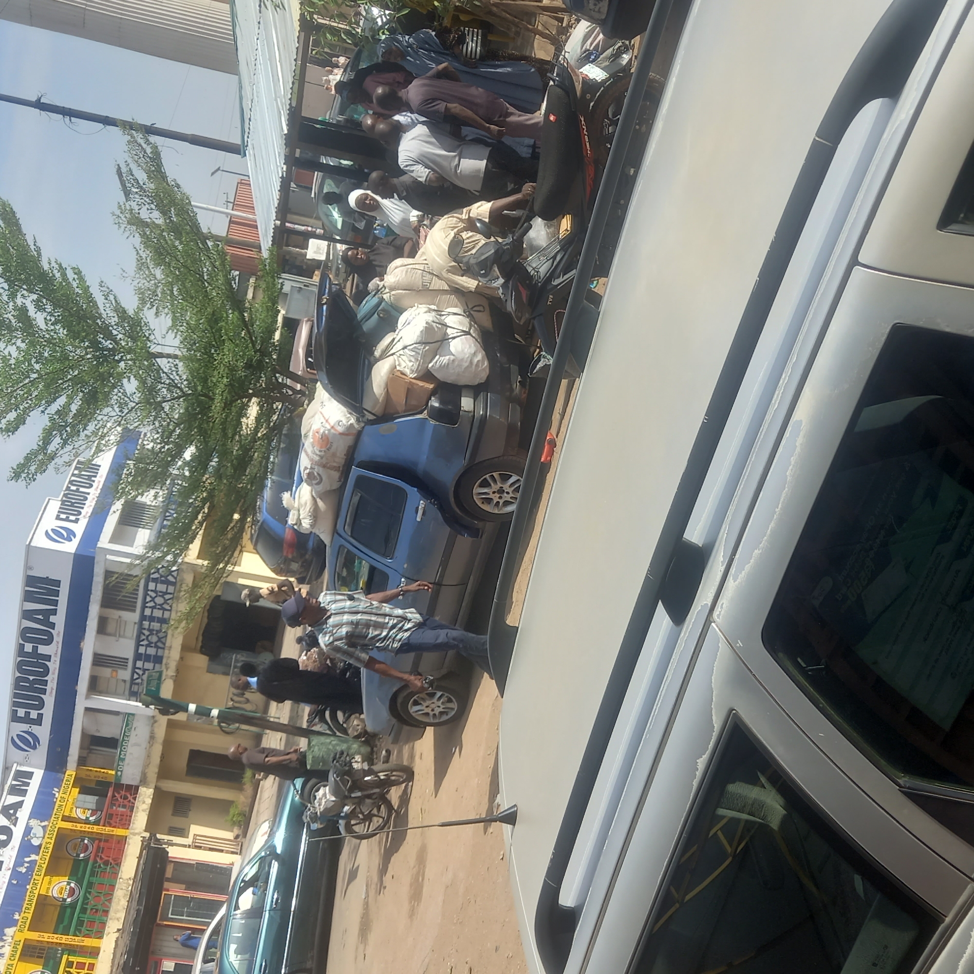 This image captures a vibrant street scene in Dadi Riba, Nigeria, characterized by human activity and vehicular traffic under bright, sunny daylight. The setting appears to be a commercial or transport area with several buildings lining the street.
In the foreground, the side of a light-colored vehicle, possibly an SUV, is visible, partially obscuring the full depth of the scene. People are the main subjects, predominantly men, engaged in various activities. One man in a plaid shirt and dark trousers stands near a light-colored car. Further back, another man is seated on a black motorcycle, heavily loaded with white sacks tied to its rear, with "TVS" visible on its side. A blue small car is conspicuous, overflowing with numerous white sacks strapped to its roof and protruding from its rear, indicating the transport of goods. Other individuals are gathered around, some wearing traditional attire, suggesting a busy communal space.
The background features green trees and several buildings. A prominent two-story building displays a large white and blue sign with "EUROFOAM" and "EUROFOAM & EUROFOAM" written on it. Adjacent to it, another building features a colorful sign bearing the text "OYA CHAPEL ROAD TRANSPORT EMPLOYER'S ASSOCIATION OF NIGERIA" and "RTEAN". Other visible text includes "ROAD SUPER", "UP ROAD", and "DE MODE" on various signs. Numbers like "0029" and "460" are also discernible, possibly associated with vehicles or street markers. The overall impression is a bustling urban environment focused on trade and transport.