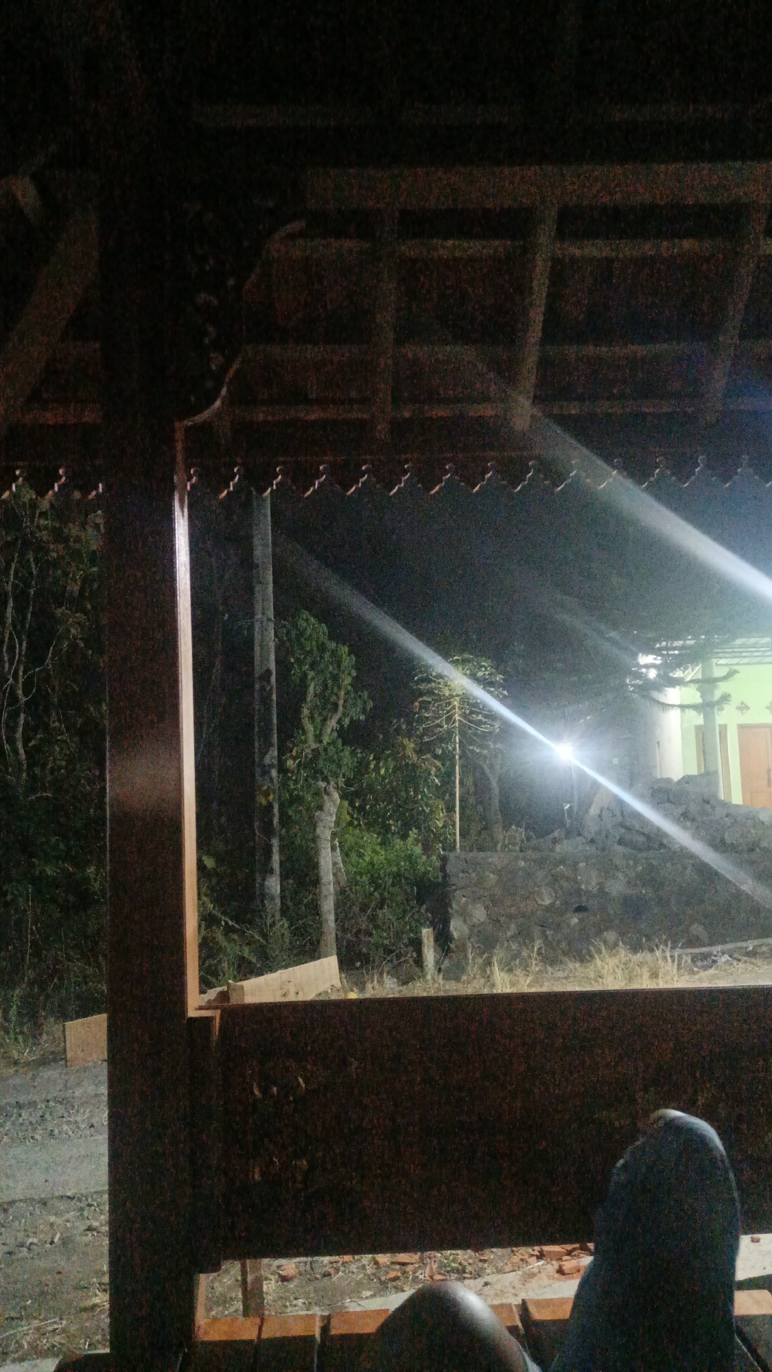 The image captures a dimly lit outdoor scene, likely at night, from within a wooden structure resembling a gazebo or veranda. The foreground is dominated by the dark wooden beams of the structure, with a section of what appears to be a wooden bench in the bottom right corner. A person's legs, clad in dark blue pants, are visible resting on this bench.
Beyond the wooden structure, the environment transitions to an outdoor setting. Trees and foliage are visible, some appearing dark and dense, suggesting a natural or rural area. A tall, slender pole, possibly a utility pole, stands prominently in the mid-ground. In the background, a building with a light-colored facade and what looks like a green roof is illuminated by a bright light source, creating strong beams of light that cut across the scene. There is a stone wall or embankment in front of the building. The overall impression is one of a quiet, possibly residential, outdoor space in the evening or at night.
Based on the provided context of Sambi, Indonesia, this scene could be a personal residence or a communal gathering spot in a village or sub-district of Sambi. The architectural style of the wooden structure and the surrounding vegetation are consistent with rural or semi-rural environments in Indonesia.