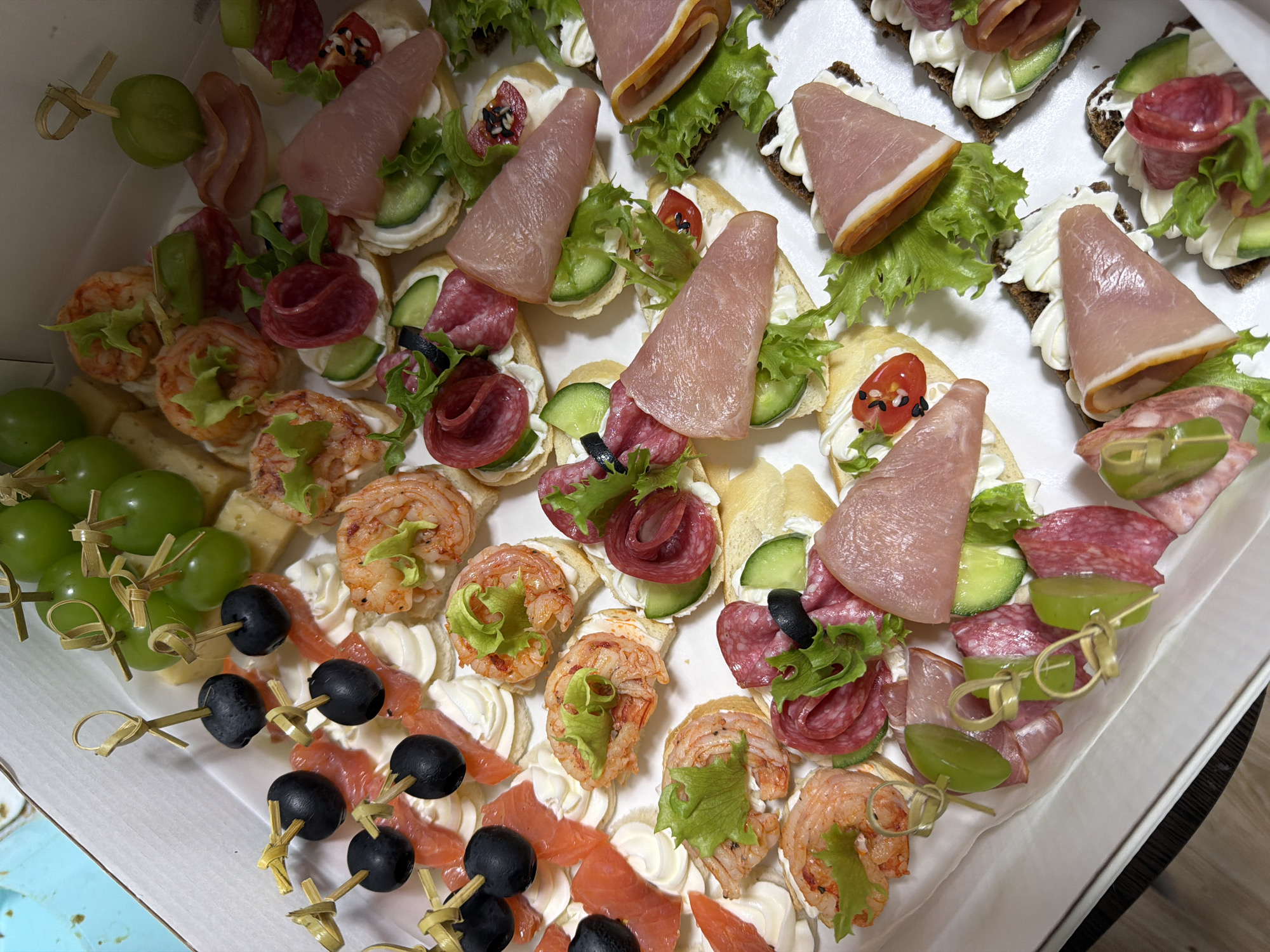 The image shows a white cardboard box filled with an assortment of appetizers. The appetizers are arranged in rows and include various small sandwiches or canapés topped with ingredients such as sliced salami, ham, shrimp, smoked salmon, olives, grapes, cucumber slices, lettuce, and what appears to be cream cheese or a similar spread. Many of the appetizers are skewered with small wooden picks, some of which are adorned with a decorative knot. The lighting suggests an indoor setting, possibly a home or a catering service. The food is presented neatly and looks ready to be served. There is no visible indication of the city of Slantsy, Russia, or any specific location information within the image itself.