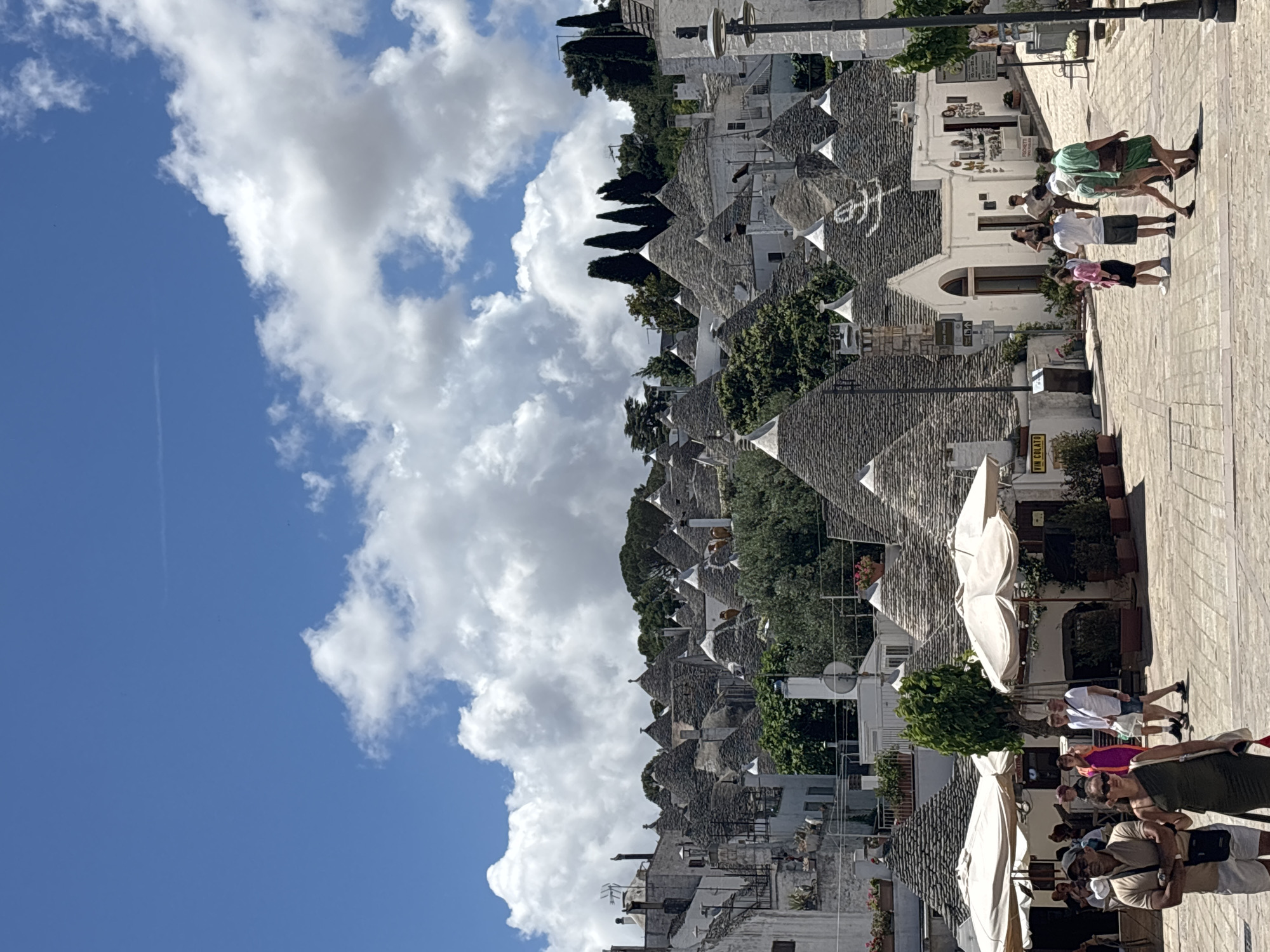 The image depicts a vibrant street scene in a town renowned for its unique architecture, likely Alberobello, Italy. Under a bright blue sky dotted with fluffy white clouds, numerous distinctive trulli houses with their characteristic conical stone roofs and whitewashed walls are densely packed, appearing to cascade down a gentle hillside. Some trulli roofs display symbolic markings.
On the right side of the image, a paved pedestrian street is bustling with people. Several individuals and small groups are seen walking leisurely, dressed in casual summer attire, suggesting a warm, sunny day. Large, light-colored market umbrellas are open along the street, providing shade over what appear to be outdoor seating areas or storefronts. Visible along the street are lampposts and commercial establishments, with one brown sign partially visible, displaying the text "VIN COLATO". The bright sunlight casts clear shadows, indicating it is likely midday or early afternoon. The overall atmosphere appears relaxed and tourist-oriented. A faint contrail is visible in the clear blue sky.