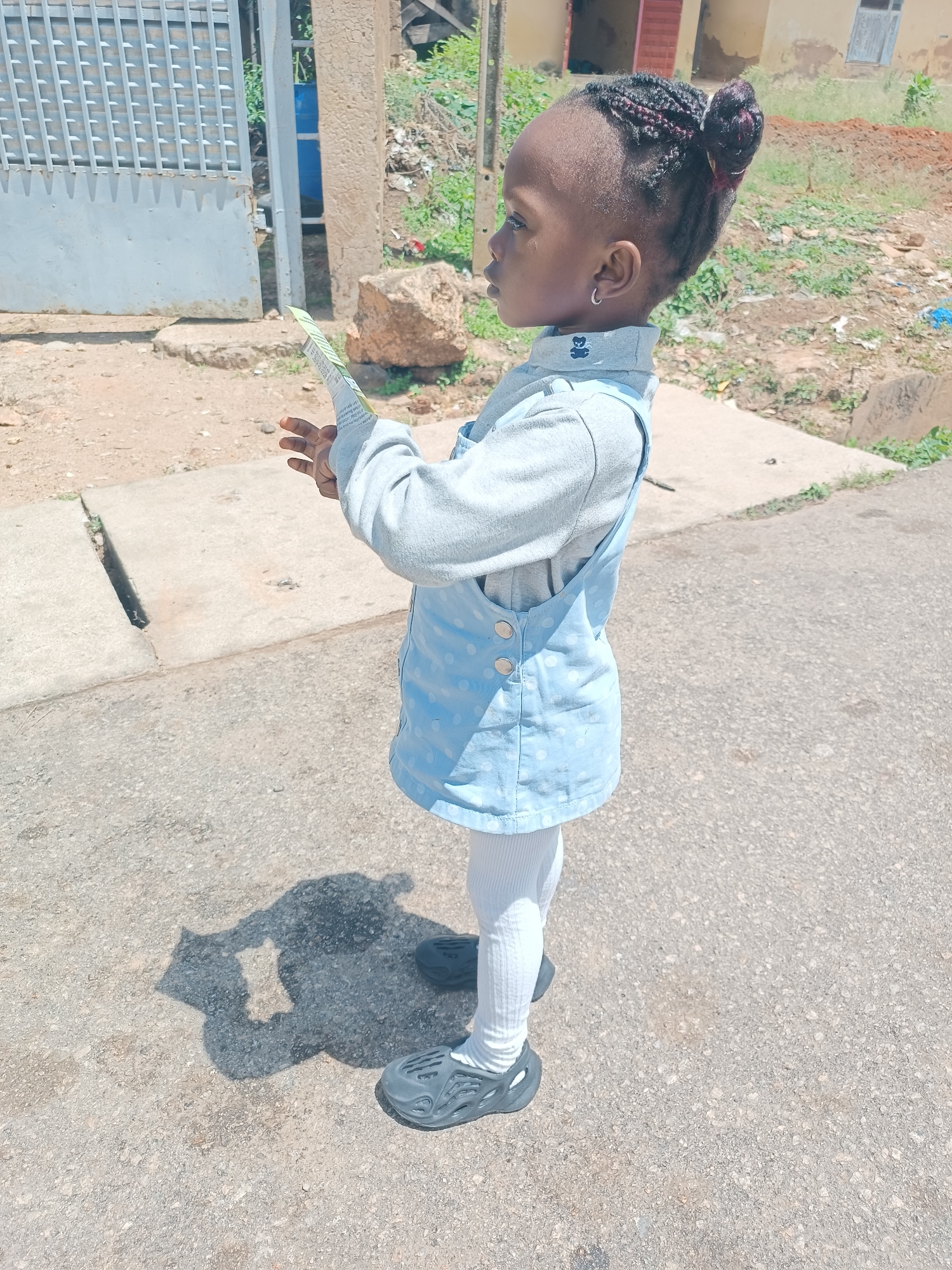 A young Black child stands on a paved road in Jos, Nigeria, holding a folded piece of paper. The child is wearing a light blue dress with white polka dots over a long-sleeved grey shirt and white ribbed tights. Dark grey clogs are on the child's feet. The child's hair is styled in braids pulled back into a bun. A silver hoop earring is visible in the child's left ear. The child's shadow is cast on the road in front of them. In the background, a grey metal gate, a blue barrel, a large rock, and buildings are visible. The environment appears to be an outdoor urban or semi-urban setting.