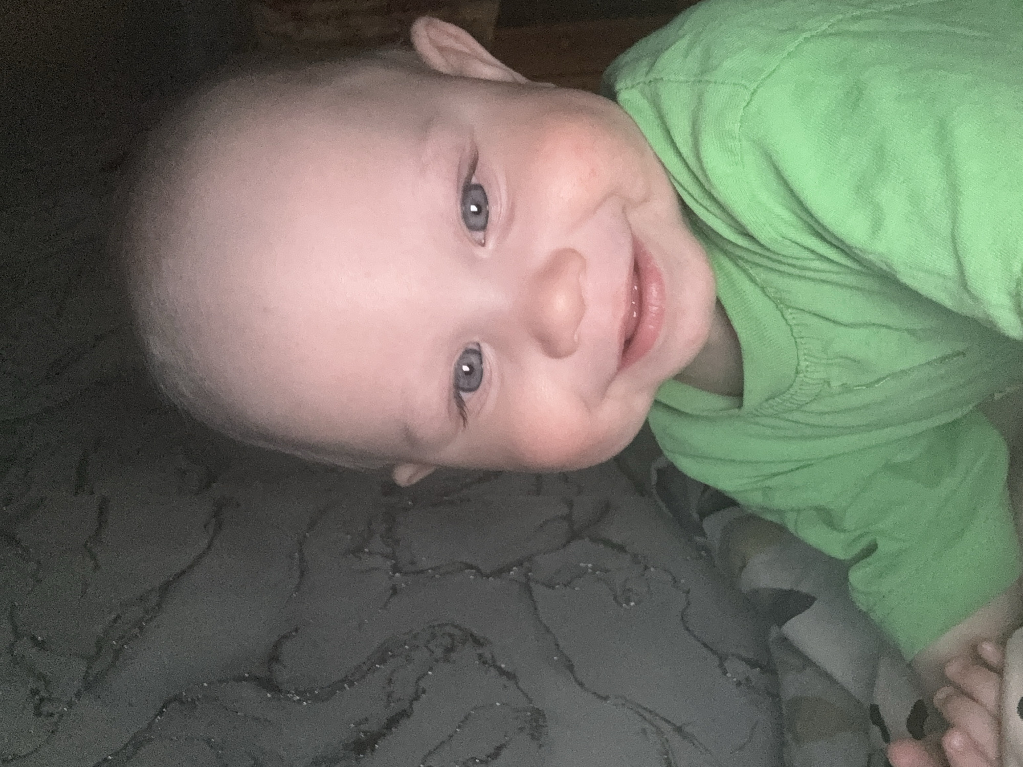 A baby lies on a textured, grey surface, possibly bedding. The baby is wearing a light green shirt and has a bald head, blue eyes, and a smiling expression. The baby's right hand is visible near the bottom right corner of the image. There is no information present in the image indicating that it was taken in the city of Chaikovsky, Russia.