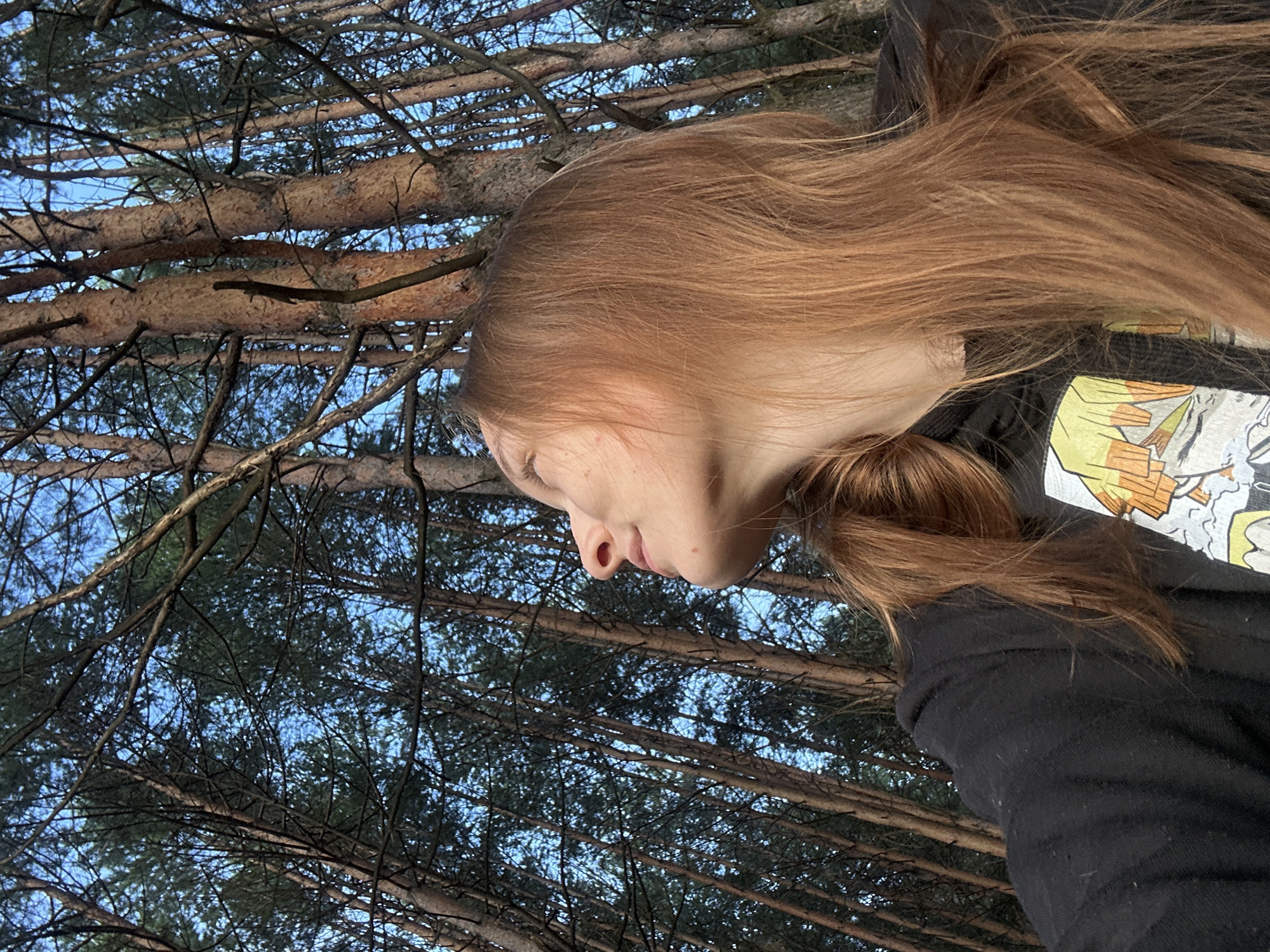 In a low-angle, medium shot taken in the forest of Chaikovsky, Russia, a young woman with long, auburn hair is looking upwards. She is wearing a black hoodie with a graphic print on the chest. The background is dominated by tall, slender pine trees and a clear blue sky. The sunlight filters through the branches, casting a warm glow on the scene. The woman's face is turned away from the camera, with her profile visible as she gazes at the sky and the trees above. Her expression appears serene and contemplative.