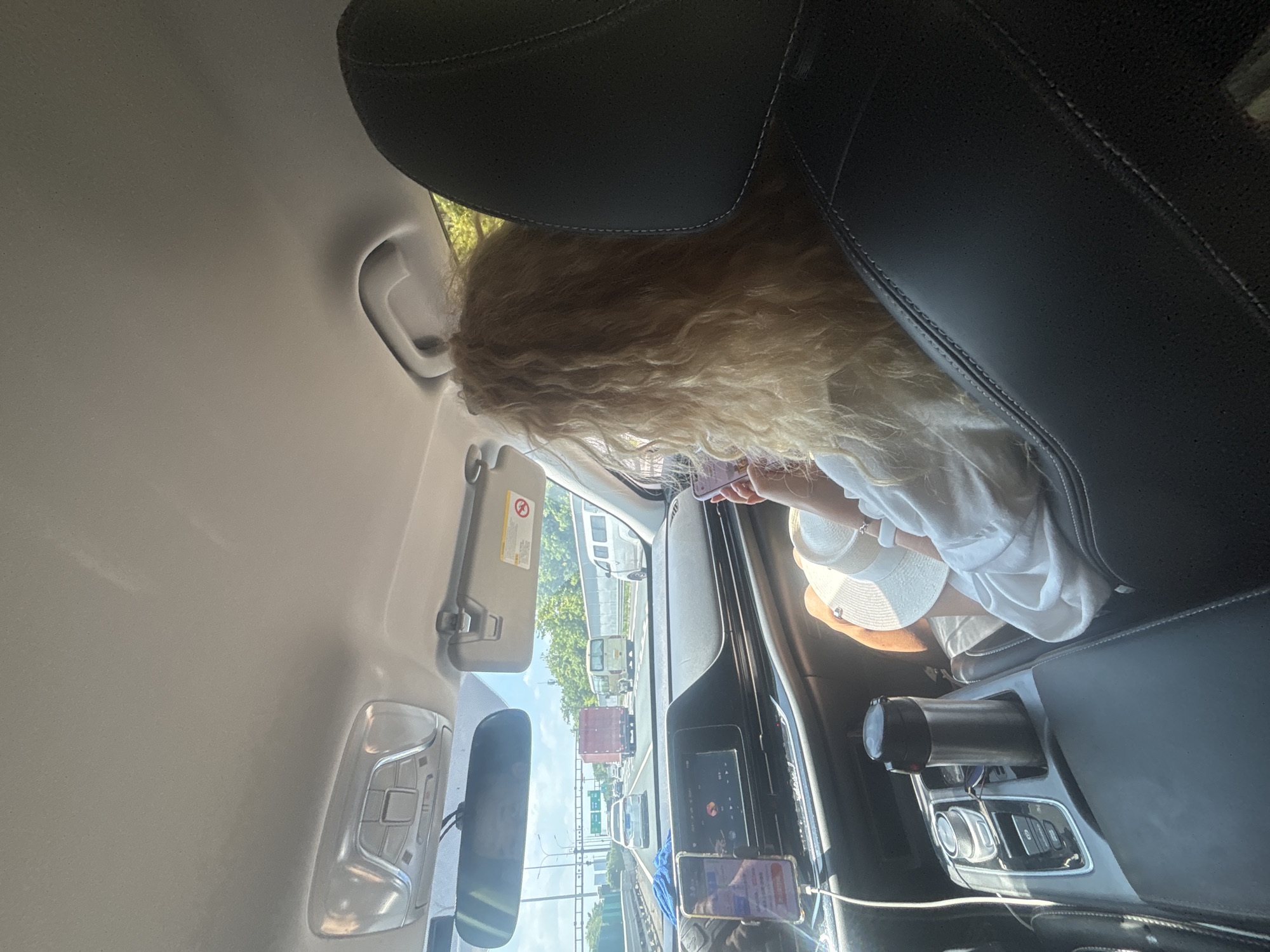The image, taken from inside a moving car, shows the passenger side of the car's interior and the road ahead. A woman with long, curly blonde hair is seated in the passenger seat, holding a white hat and a smartphone. Her left arm is resting on the center console. The car's dashboard is visible, including a large infotainment screen and a phone mount with a smartphone displaying a navigation app. A thermal flask is situated in a cup holder on the center console. The road ahead is a multi-lane highway with other vehicles, including trucks and cars. Trees and a sound barrier are visible along the roadside. The sky is clear and bright. The scene appears to be in the Liwan District, China, indicated by the car's navigation system displaying Chinese characters.