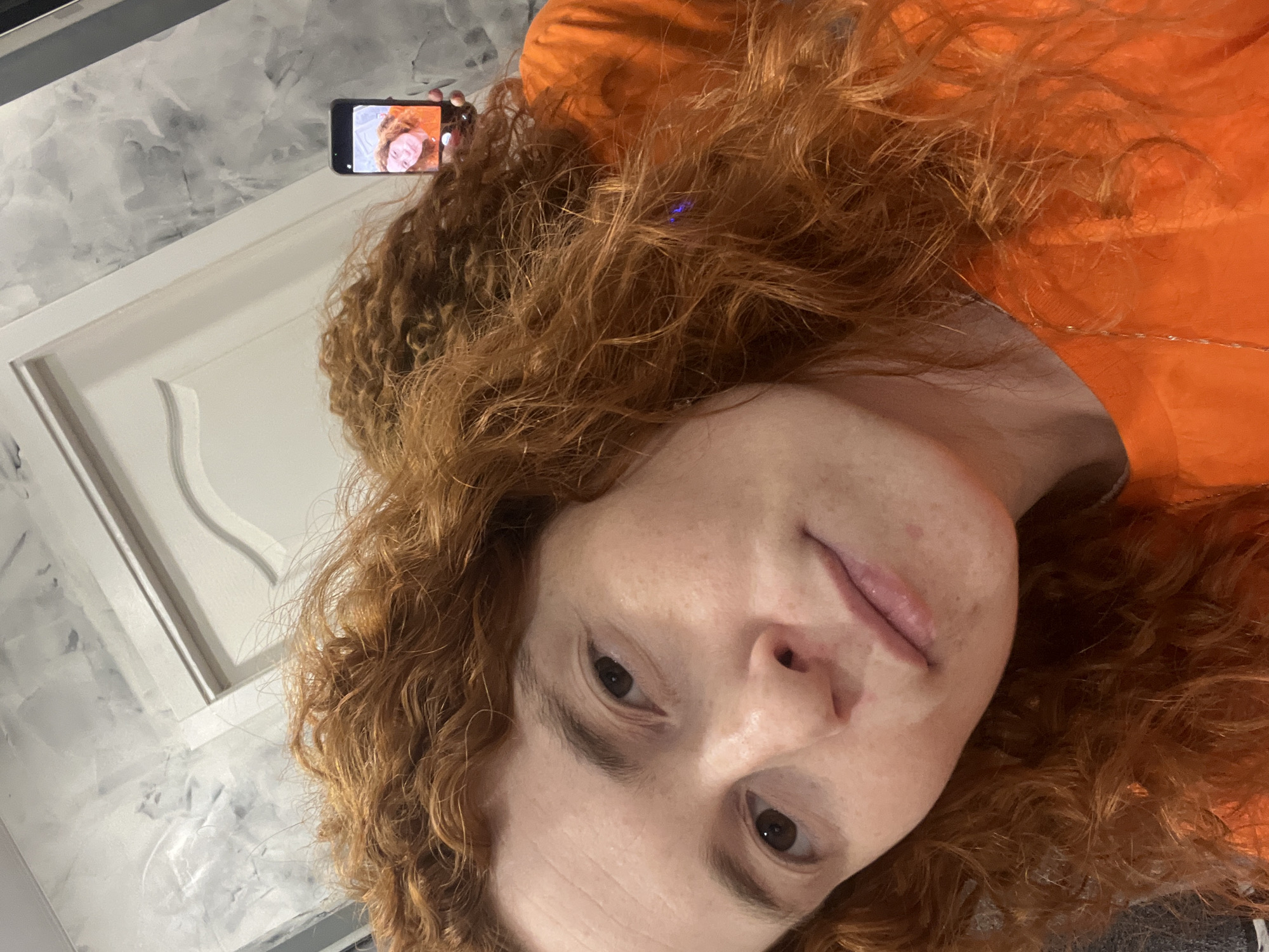 A close-up, rotated image captures a person taking a selfie, creating a "photo within a photo" effect. The central subject is a person with fair skin, dark eyes, and abundant, curly, reddish-brown hair. They wear an orange top or shirt. The person gazes directly towards the camera, with a neutral to slightly contemplative expression.
In the upper left background, the person is holding a black smartphone. The screen of this phone is clearly visible, displaying a live preview of the person's face, which appears upside down and mirrored relative to the main subject. This suggests the primary image is a photograph of a person taking a selfie, possibly reflected in a mirror or captured by another camera, rather than the selfie itself.
The background features a light-colored wall with a textured, mottled, or marbled finish. A white door frame or paneling with decorative molding is visible on the left. The scene is indoors and well-lit. The overall image is rotated 90 degrees clockwise. There is no visible text. The setting is an interior in Chaikovsky, Russia.