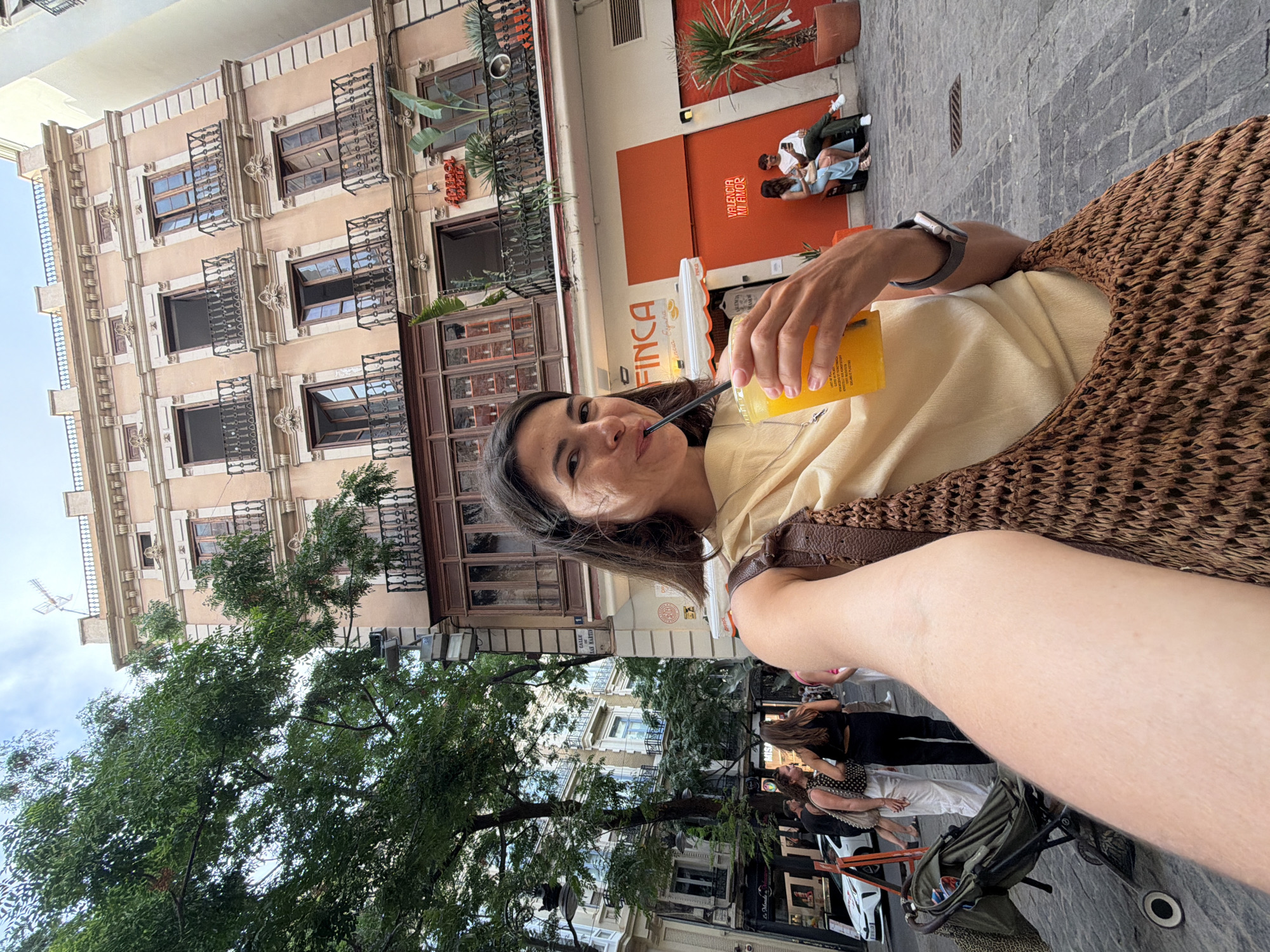A woman is taking a selfie in an urban setting. She is holding a plastic cup of orange beverage and drinking it through a dark straw. She is wearing a light yellow sleeveless top and a woven brown bag is slung over her shoulder. The background shows a pedestrian street with buildings and trees. One building has balconies and ornate architecture. Another building, painted orange, has a sign that reads "VALENCIA MI AMOR". Two people are sitting on a bench outside the orange building, looking at their phones. A stroller is visible in the foreground to the left. The ground is paved with cobblestones. The sky is visible at the top of the image.