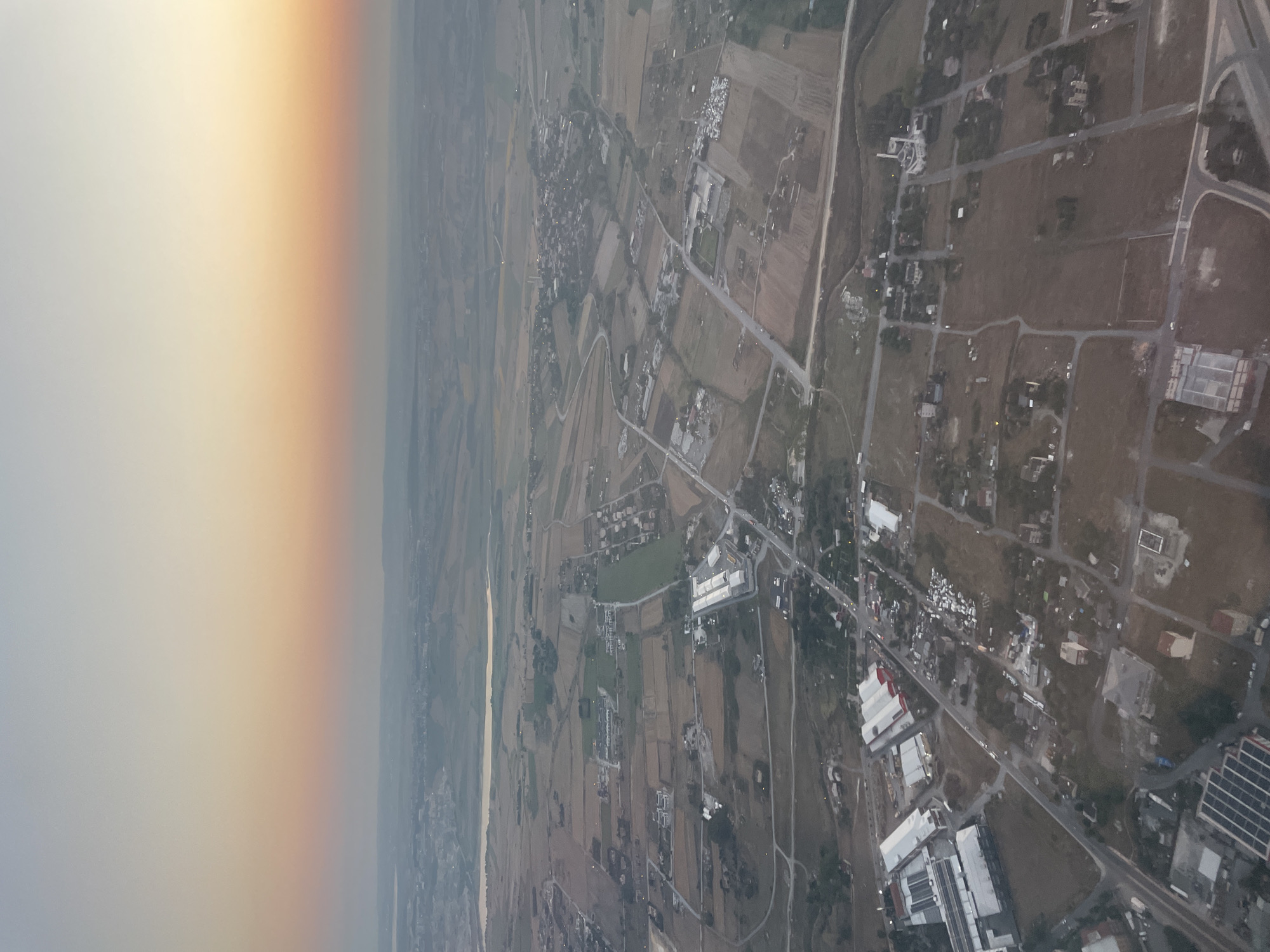 An aerial photograph captures a sprawling urban landscape under a hazy, gradient sky transitioning from pale blue to orange. The scene depicts a mix of agricultural fields, residential areas, and industrial structures, with a notable presence of a mosque with a prominent dome and minaret visible in the middle ground. Roads crisscross the terrain, connecting various buildings and parcels of land. In the foreground, a cluster of industrial buildings with white roofs and red accents is apparent. Further in the distance, a body of water can be observed. The overall impression is of a developing area on the outskirts of a city, with evidence of both urban expansion and existing rural land use.