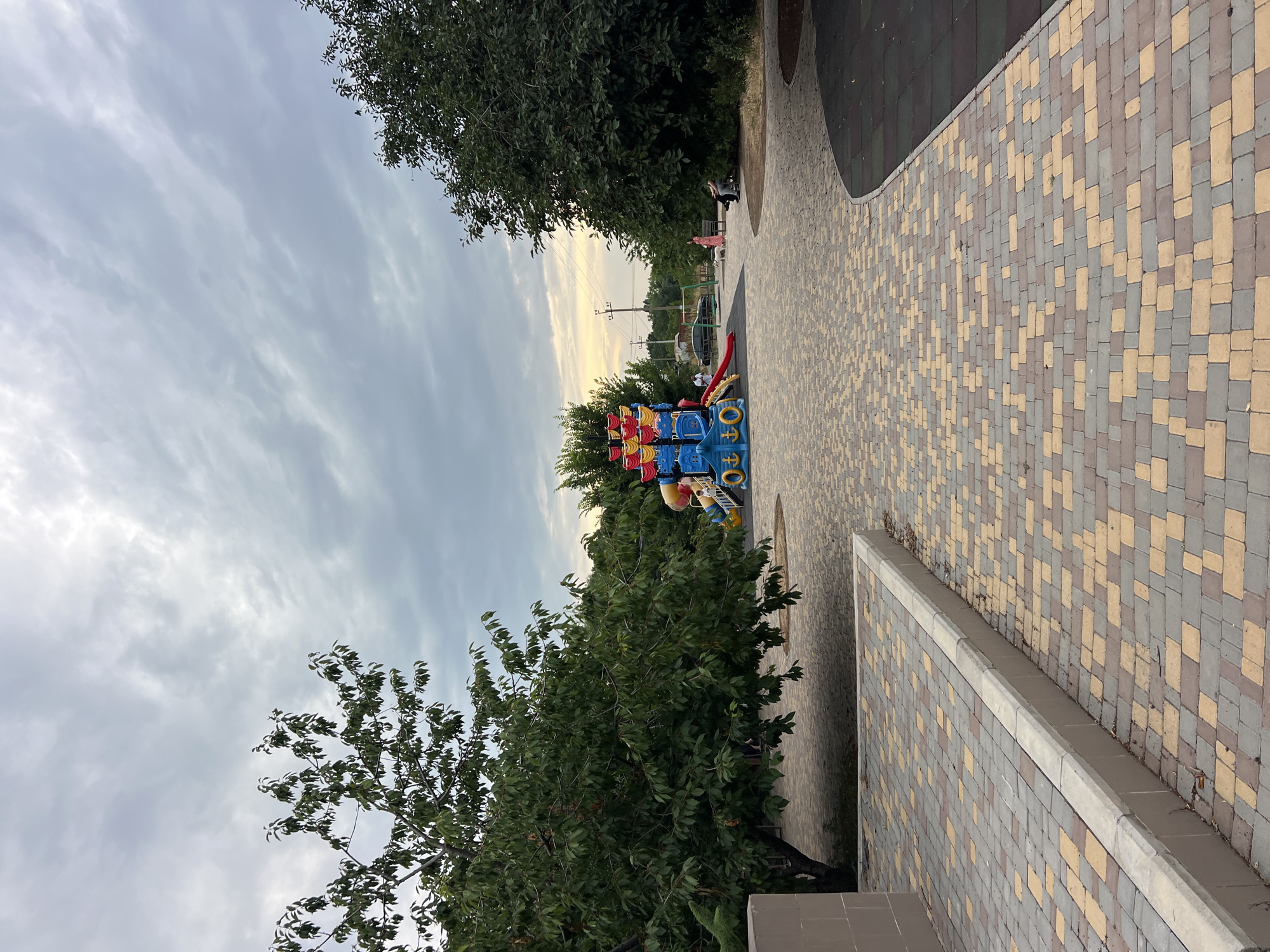 In Fontanka, Ukraine, an outdoor paved area features a colorful playground structure resembling a pirate ship, complete with slides and climbing elements. The ground surrounding the playground is a mix of rubberized safety surfacing and patterned brickwork. Adjacent to this area, a large tree with dense green foliage provides shade. Further away, a few individuals are visible; two people sit on a bench, while another stands near a swing set. The sky overhead is overcast with gray clouds, and power lines are visible in the distance. The overall scene suggests a public park or recreational space.