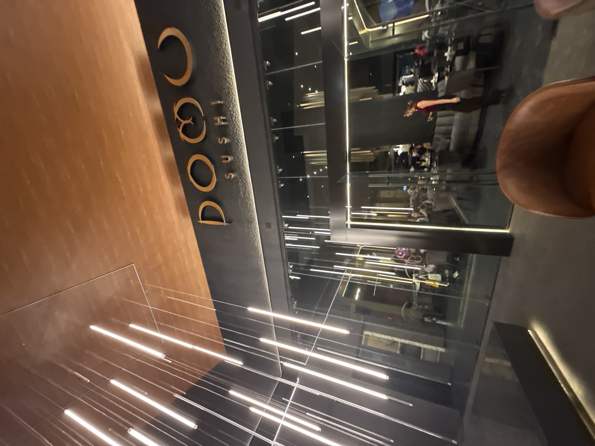 The image depicts the interior of a restaurant named "DOGO Sushi." The signage is in gold lettering on a dark background. There are numerous linear light fixtures extending from the ceiling. Through the glass walls, blurred figures of people can be seen inside the restaurant. A brown chair is visible in the foreground. The restaurant is located in San Bernardino Tlaxcalancingo, Mexico.
