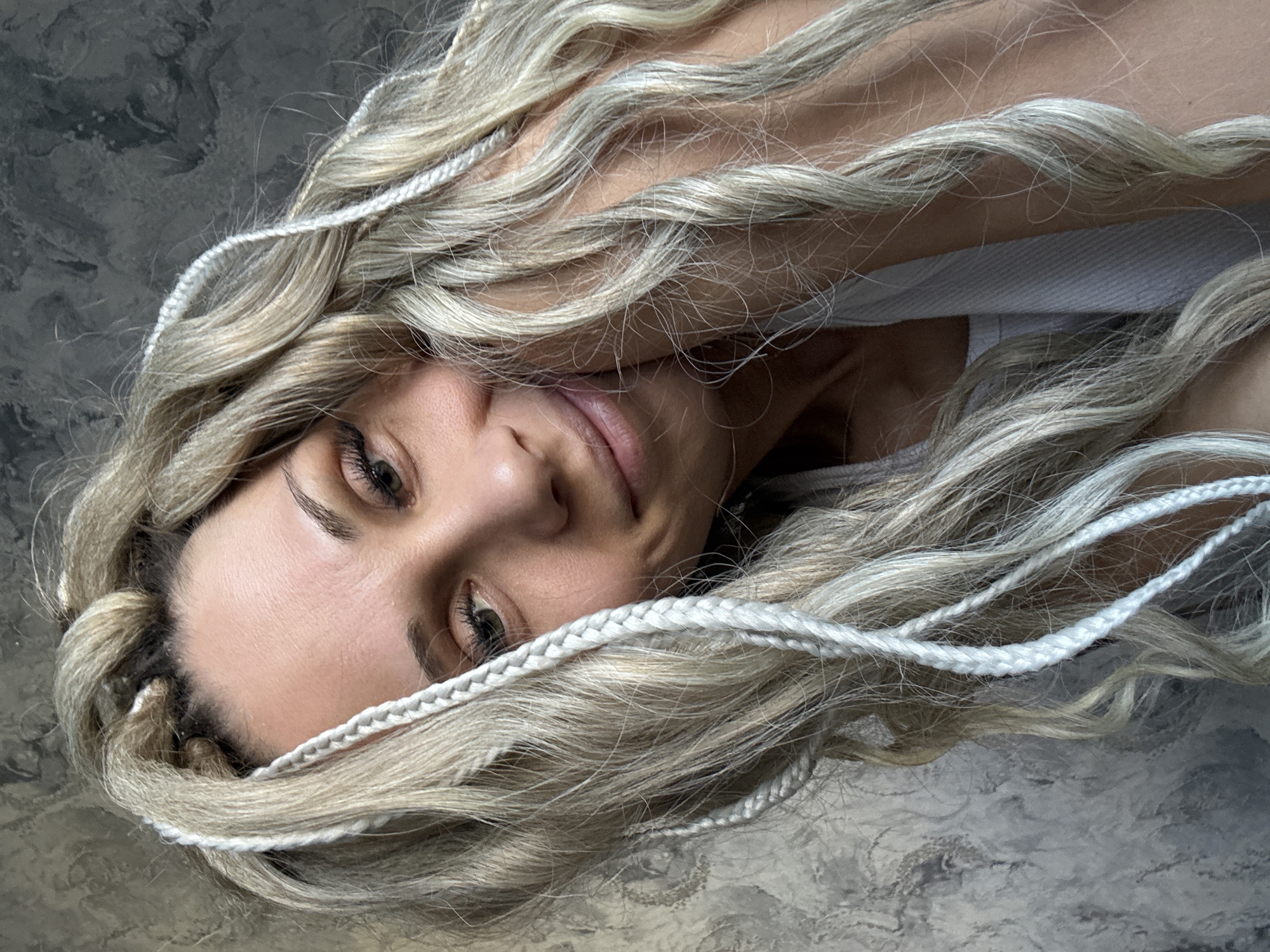 A fair-skinned woman with blond hair lies prone against a mottled gray surface. Her hair is styled with numerous thin, white braids interwoven. She wears a white top. The location is not discernible as Rusanovo, Russia.
