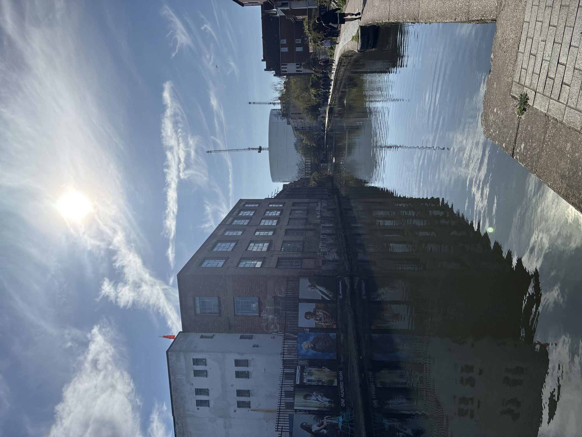 An eye-level, landscape-oriented shot shows a waterway reflecting several buildings, sky, and the sun. On the right of the water is a paved sidewalk. The buildings reflected in the water are predominantly brick and white. On the brick building are posters of people. Two cranes are visible in the background. The sky is blue with wispy clouds. The sun is positioned in the upper-left corner of the frame. It appears to be a canal in the London area of the United Kingdom. A person is walking on the sidewalk to the right of the canal.