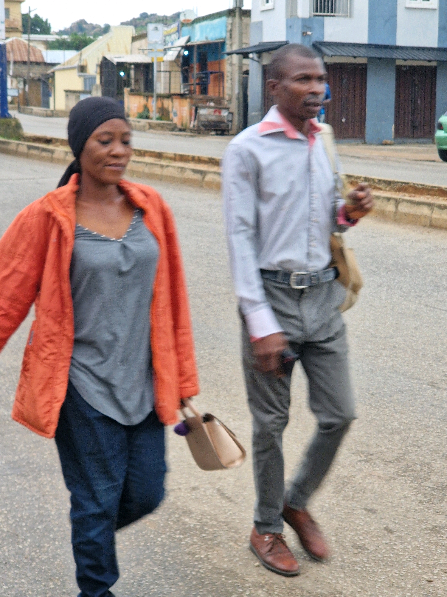 Two individuals, a woman and a man, are walking on a paved street in an urban environment. The woman, on the left, wears a black headscarf, an orange jacket, and a grey top with a v-neck and metallic detailing around the neckline. She also has on dark blue jeans and is carrying a light brown handbag with a dark purple pom-pom attached. The man, on the right, is wearing a light blue button-down shirt with a pink collar, grey pants, a dark belt with a silver buckle, and brown dress shoes. He has a light brown bag slung over his right shoulder. In the background, buildings with various colors and architectural styles line the street, suggesting a commercial or residential area. A portion of a green vehicle is visible on the far right of the frame. The ground appears to be asphalt with some visible texture. The lighting suggests it is daytime, possibly overcast.