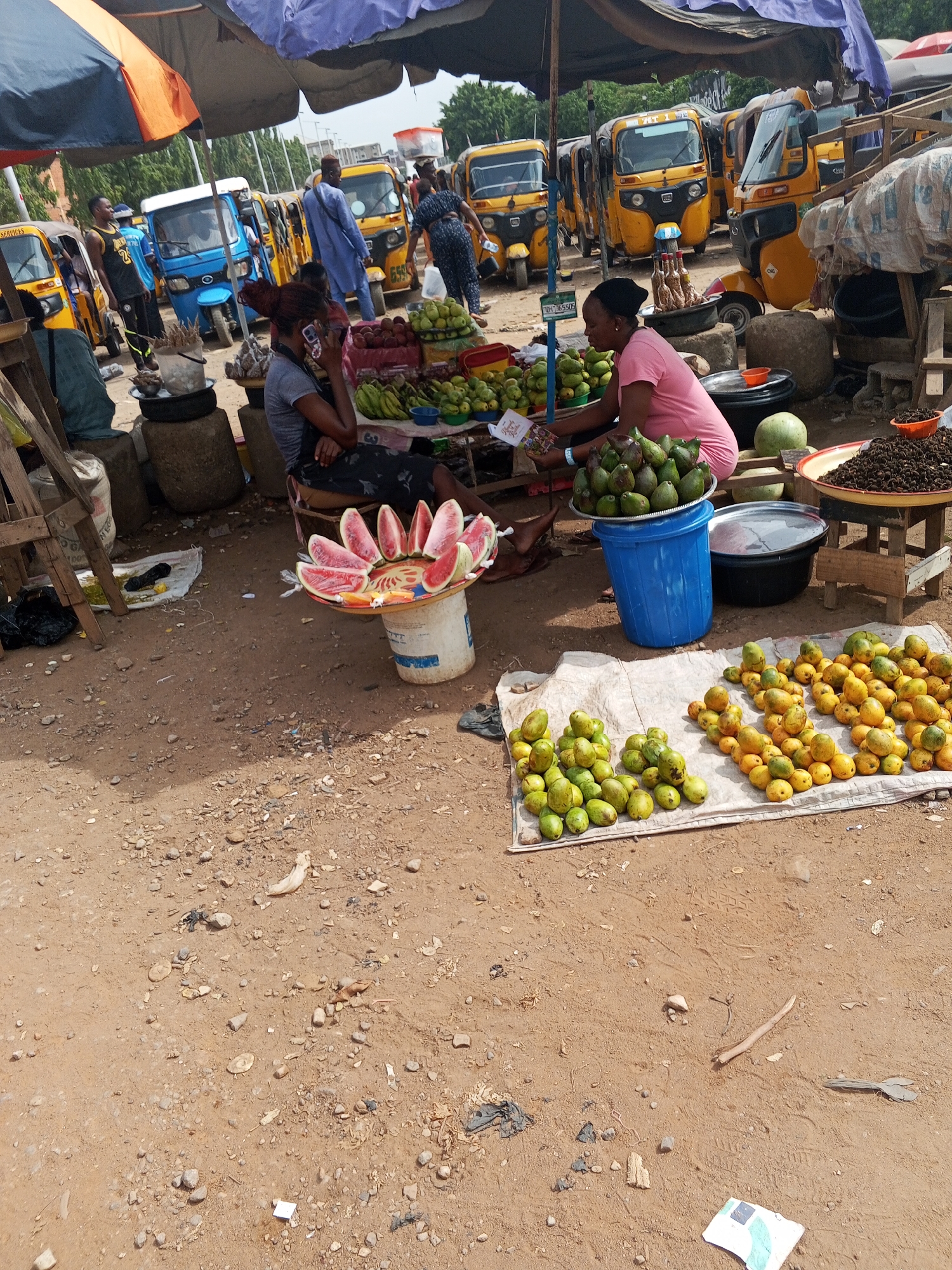 The image depicts a bustling roadside market in Abuja, Nigeria. Two women are seated under a makeshift shade selling a variety of fruits: sliced watermelon, avocados, mangoes, bananas, and other local produce. One woman is on her phone. The produce is displayed on tables, buckets, and a cloth spread on the ground. Several yellow and blue auto-rickshaws (tricycles) are parked in the background, suggesting a transportation hub. Other people are visible, some walking around and others near the auto-rickshaws. The ground is dirt with scattered debris. Overhead, a colorful umbrella and blue tarp provide shade.