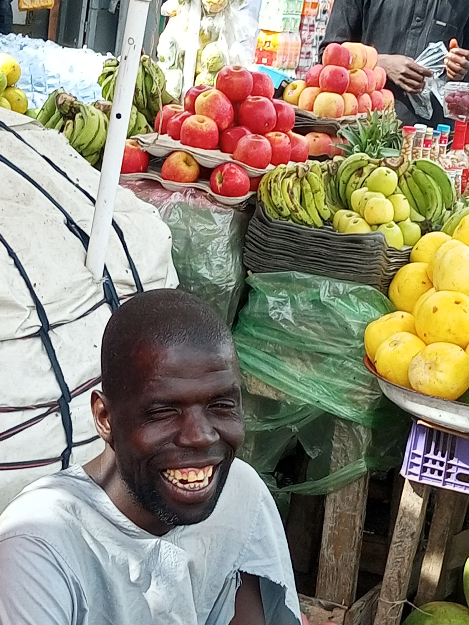 A man is smiling in a market. He is wearing a light blue t-shirt. His head is turned to his right, and he is winking with his left eye. His mouth is open in a wide smile, revealing his teeth. He has short, dark hair and a beard.
Behind the man, there is a variety of fruits for sale, including bananas, apples, and what appear to be guavas or a similar citrus fruit. The fruits are displayed on tables and in baskets. Some of the fruit is wrapped in plastic. A white pole is visible in the middle ground, and a white tarp is tied with black ropes behind the man. In the background, there are shelves filled with bottles and jars. The scene appears to be outdoors, likely in a market or street vendor setting. The lighting suggests it is daytime, possibly morning or afternoon. The overall atmosphere is cheerful, due to the man's expression. No visible text is present in the image.