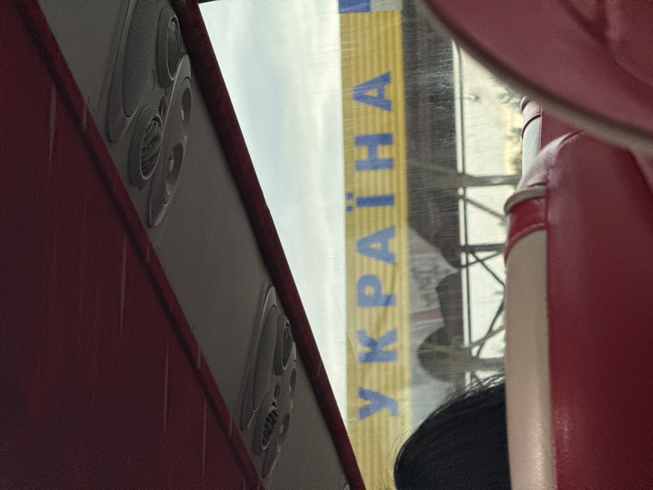 The image depicts a scene viewed from inside a vehicle, likely a bus, looking outward through a window. The upper section of the vehicle's interior is visible, featuring a textured red ceiling and a central overhead panel in a lighter gray shade, equipped with multiple circular air vents and integrated light fixtures. The lower portion of the frame shows red-upholstered seating, with a headrest and a portion of a seat prominently featured. The back of an individual's head, displaying dark hair, is partially visible on the left side, suggesting a passenger in the seat immediately in front of the camera's position.
Through the vehicle's window, an exterior scene is visible, dominated by a large yellow banner emblazoned with bold blue Cyrillic letters spelling "УКРАЇНА" (UKRAINE). This banner is mounted on a structural framework, indicating a border crossing facility. The sky beyond the banner appears light and overcast. The specific location is the Starovoitove border crossing in Ukraine, where the sign welcomes entry into the country. Faint, indistinct vegetation is visible in the distance below the main sign structure.