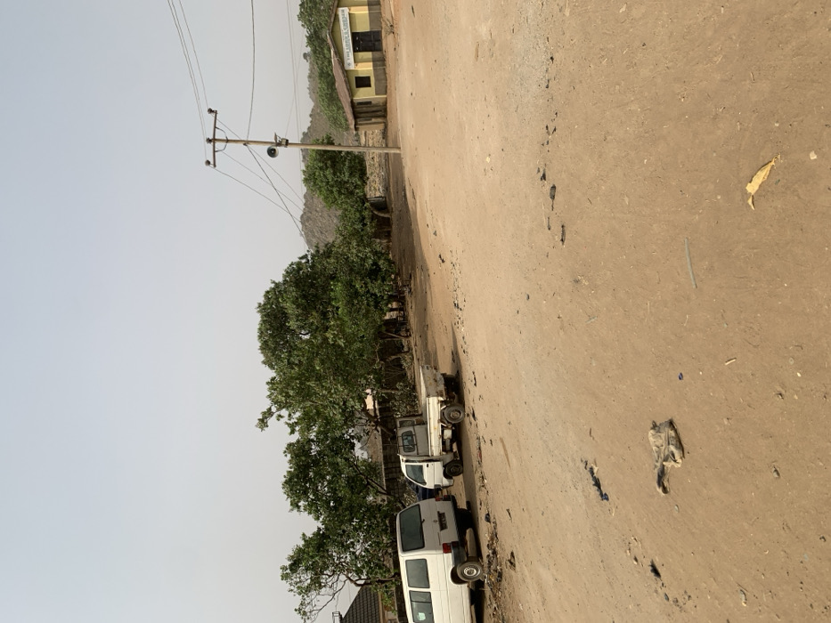 This image depicts an unpaved, dusty road scene in Bwari, Nigeria, under a pale, hazy sky. The foreground and mid-ground consist of reddish-brown dirt, with scattered refuse including a light-colored fabric item, a yellow leaf, and a blue plastic cap. On the left, a white passenger van is parked, followed by a smaller white pickup truck behind it. A cluster of green-leafed trees and the upper portion of a building's roof are visible on the far left. To the right, a single-story building with yellow walls and a brown base displays a sign reading 'THE LORD'S CHOSEN'. A wooden utility pole, equipped with multiple overhead wires and a loudspeaker, stands near the center-right, positioned in front of this building. The background reveals rocky outcrops and additional green trees. No individuals are visible, and no discernible actions or interactions are occurring within the frame.