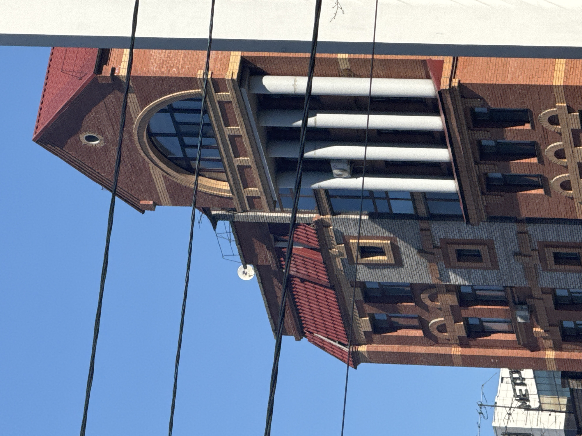 The image shows an architectural detail of a brick building against a blue sky, with power lines visible in the foreground. The structure features a combination of red brick and gray brick, along with prominent design elements such as arched windows, what appear to be ventilation shafts, and decorative brickwork. Part of a white sign with text is visible below the building.
Based on visual cues, the building in the picture is likely the Krasnoyarsk Regional Science Library located in the city of Krasnoyarsk, Russia.