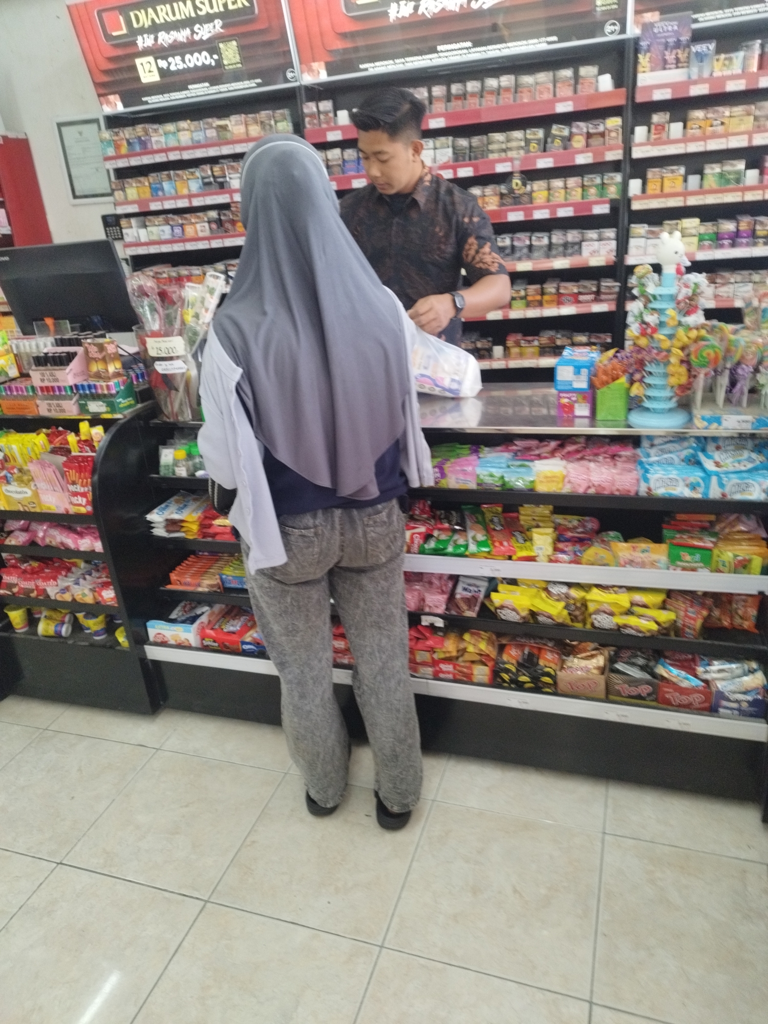 In Kepoh, Indonesia, a person wearing a gray hijab and gray patterned pants stands with their back to the camera in a convenience store. They are looking at a shelf of snacks, with a male store employee in a dark patterned shirt visible behind them, looking down. The store shelves are stocked with various food items, including cigarettes, snacks, and candy. The floor is tiled.
