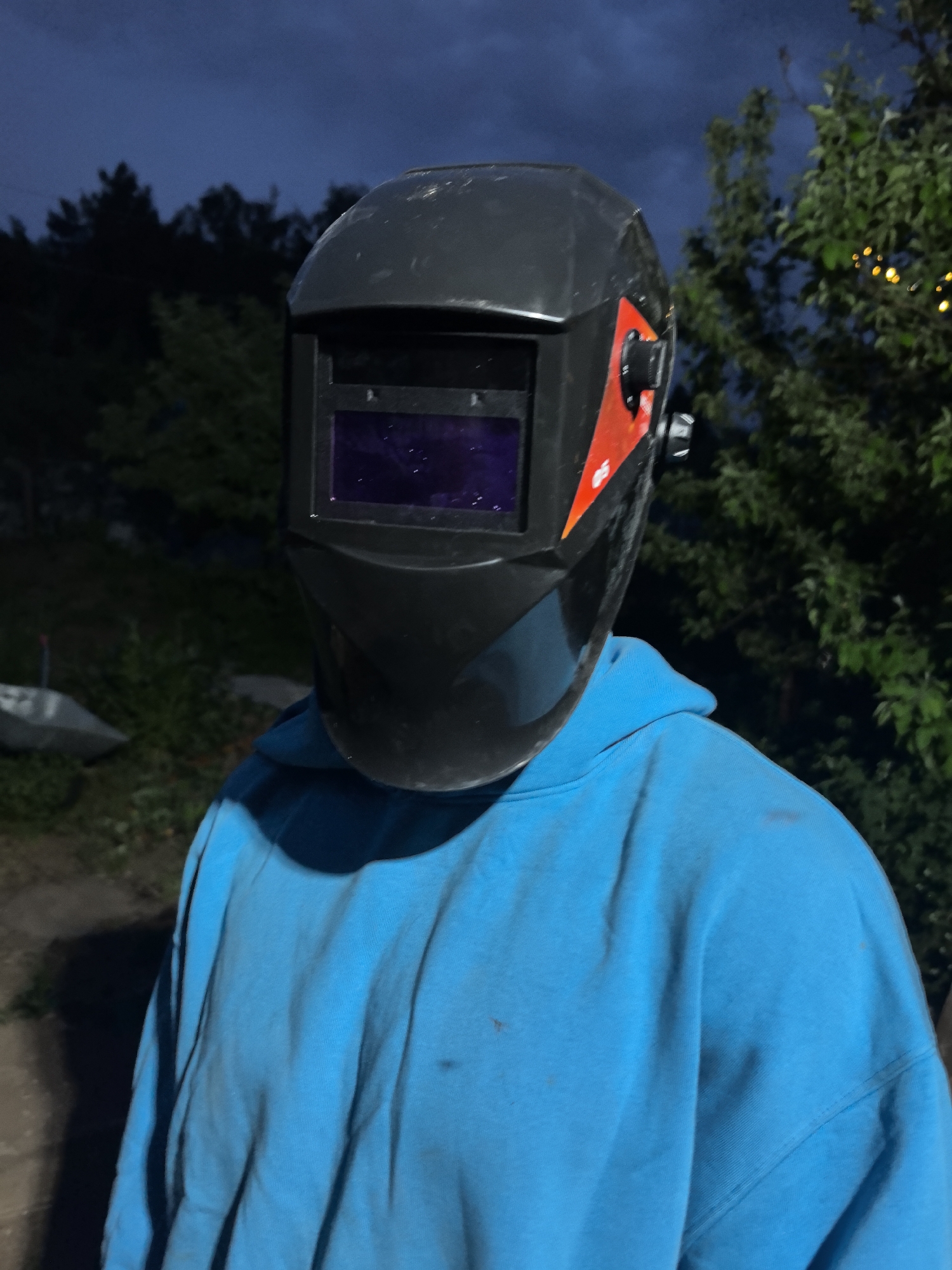 A person wearing a black welding helmet and a bright blue hoodie stands outdoors. The background features trees and a dark sky. The person's face is obscured by the helmet. There is a black knob and red accents on the side of the helmet. The location is outside, likely in a yard or natural area, with a blurred background. The hoodie appears to be worn and possibly stained. There is no visible sign of the city of Noviy, Russia, in the picture.