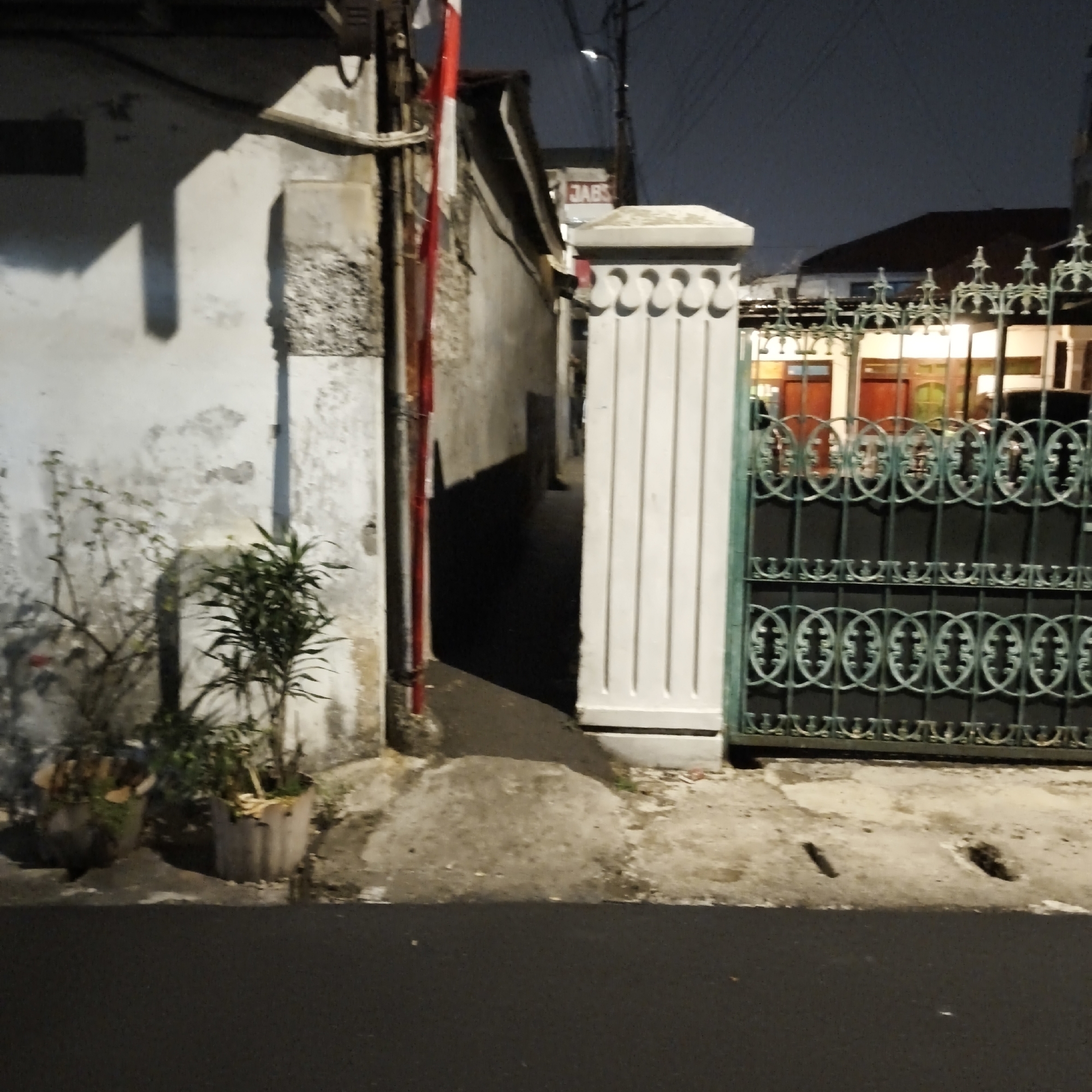 The image captures a nighttime scene in South Jakarta, Indonesia, focusing on an alleyway entrance leading to a property. To the left, a weathered white wall is partially illuminated, showing signs of age and a downspout. Two potted plants are situated near the wall, one with sparse foliage and the other a denser, green plant. The alleyway itself is a dark, paved passage, with a strong shadow cast by unseen structures.
On the right side of the alley, a white ornamental pillar marks the entrance. Beyond the pillar, a green wrought-iron gate is visible, adorned with decorative circular patterns and a more intricate top railing. Through the gate, a dimly lit building facade is discernible, with red-toned doors or windows. A sign with the letters "JABS" is mounted on a building further back, above the gate. Overhead, electrical wires crisscross against the dark night sky. The ground in the foreground is asphalt, contrasting with the textured concrete and paving stones of the alley and walkway. The overall impression is one of a residential area at night, with a focus on the entrance and boundary of a property.