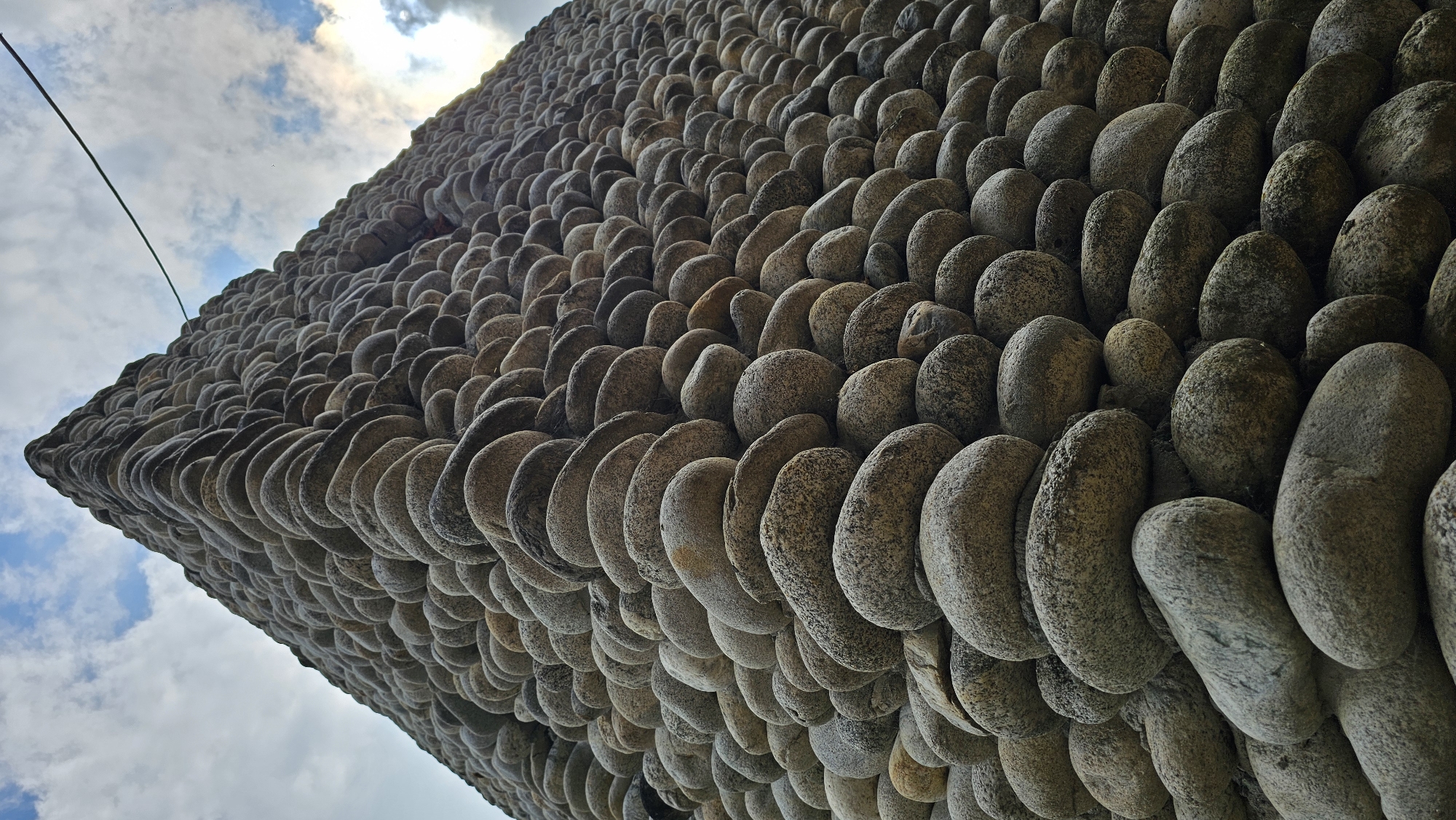 The image shows a low-angle, upward-looking view of a tall, triangular structure constructed entirely of smooth, rounded stones. The stones are of varying sizes and shades of gray and brown, and are meticulously stacked to form the sides of the structure. The perspective emphasizes the height and scale of the construction, with the peak disappearing into a cloudy sky. A thin black wire or cable is visible stretching diagonally across the upper right portion of the frame, against the sky. The overall impression is one of a unique architectural or artistic creation, likely an outdoor sculpture or building element. The image does not depict any people, specific actions, or interactions. The location is identified as the city of Krasnyy Kurgan, Russia, implying this structure is situated within an urban or semi-urban environment.