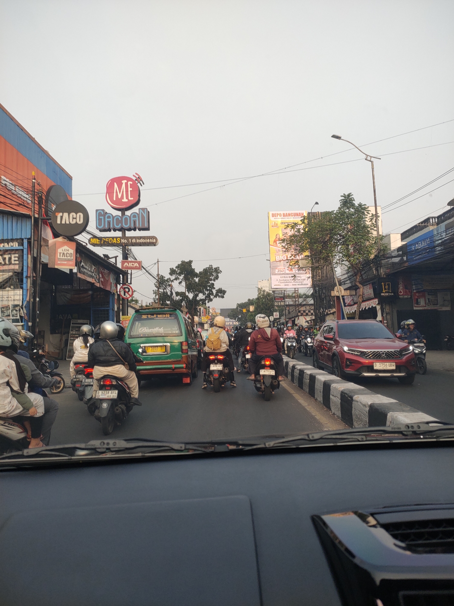 The image shows a street scene in Cimahi, Indonesia, with heavy traffic consisting of motorcycles and cars. On the left side of the street, there are several buildings with signs for "Taco" and "ME Gacoan" which appears to be a restaurant. The buildings have a distinctive orange and blue color scheme. A green van is in the middle of the road, followed by a red SUV. Several people on motorcycles are visible, some wearing helmets and casual clothing. The sky is overcast. The image is taken from the perspective of someone inside a car, looking forward through the windshield. The road is divided by a concrete barrier with black and white markings. Advertising billboards are visible in the background.