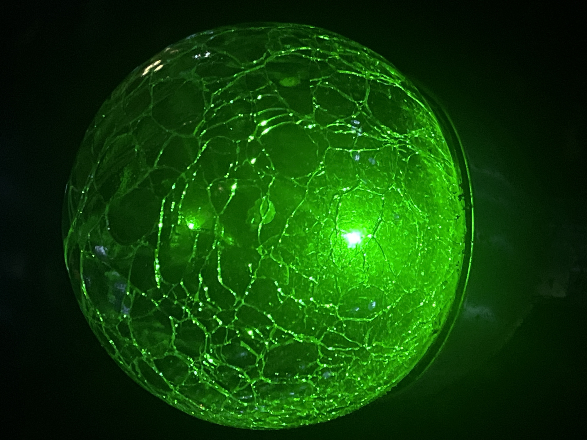 A cracked glass sphere, illuminated internally with a bright green light, is positioned in the center of the frame against a dark background. The sphere's surface is covered in a network of fine cracks, resembling a shattered mosaic, with the light shining through these fissures, creating a luminescent, web-like pattern. A brighter green light emanates from the lower center of the sphere, indicating the primary light source within. The overall impression is one of an otherworldly or decorative object, its material properties enhanced by the internal illumination. There is no indication of location in Branipole, Bulgaria, or any other specific environmental context.