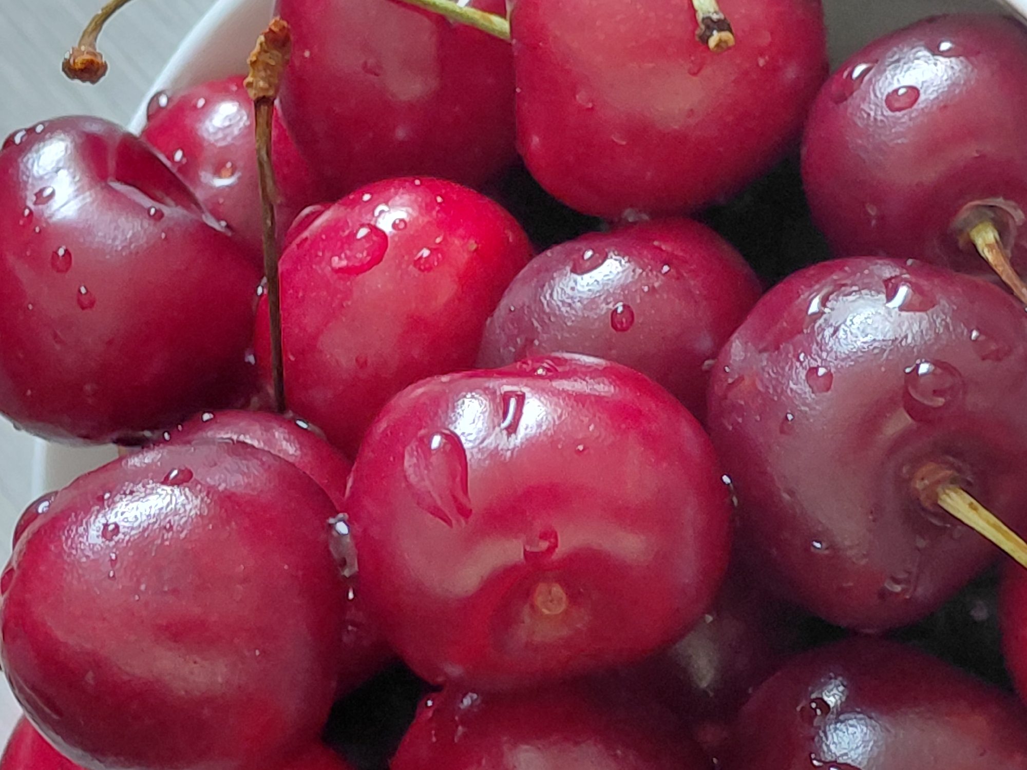 The image displays a close-up, overhead view of a bowl filled with fresh, ripe cherries. The cherries are a deep red color and appear to be glistening with water droplets. Some cherries have stems attached, while others do not. The lighting suggests an indoor setting, possibly a kitchen or dining area. The background is blurred and appears to be a light-colored surface, possibly a table or countertop. There are no people or specific landmarks visible to indicate a location within Novosibirsk, Russia. The scene is a still life focusing on the natural beauty and freshness of the fruit.