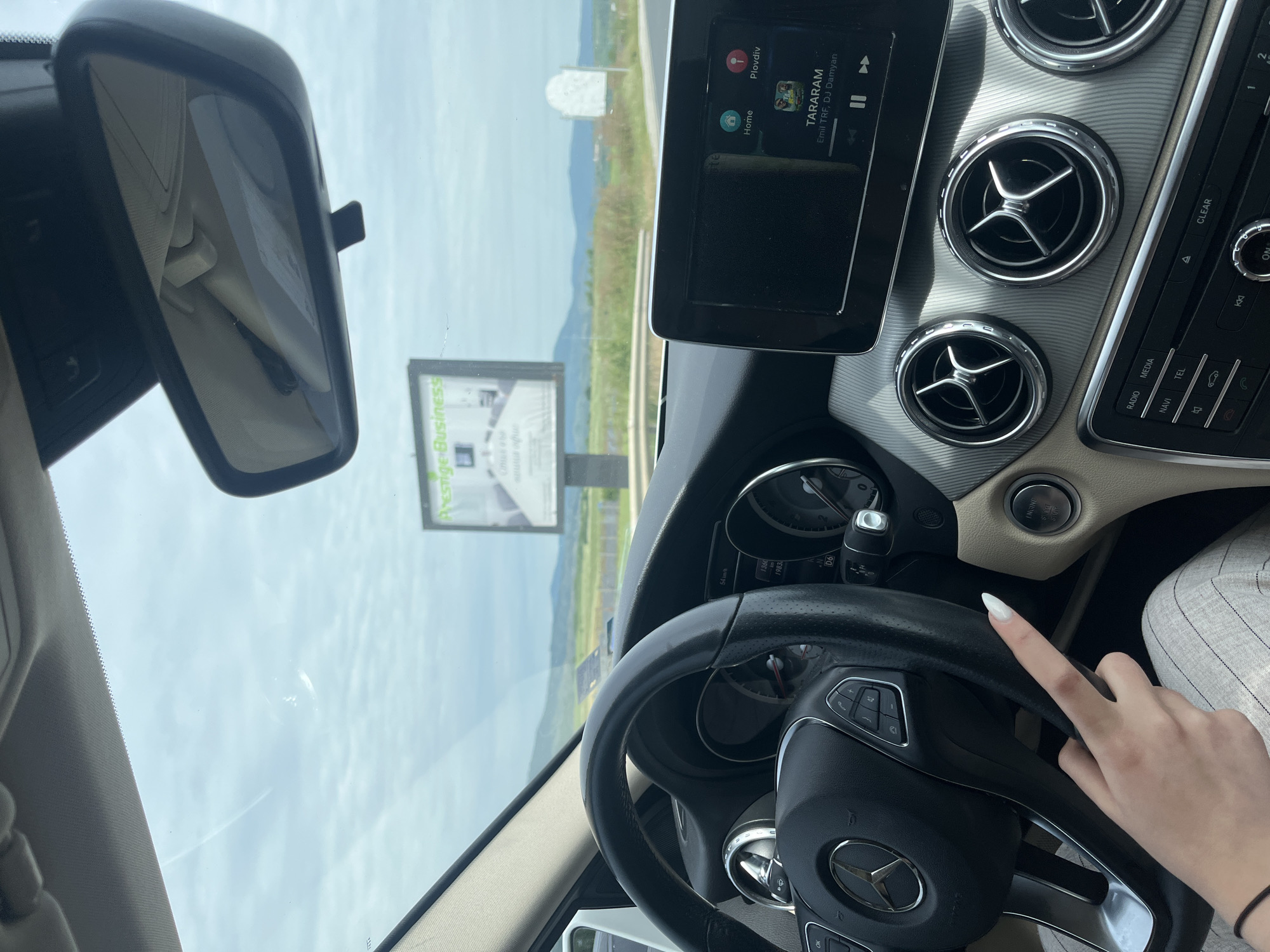 The image shows a view from inside a car, with a dashboard and steering wheel prominently featured. The steering wheel has a Mercedes-Benz logo. A hand with long white nails is visible, near the steering wheel. The car's infotainment system is displaying the song "TARARAM" by Emil TRF, DJ Damyan. Outside the car, a billboard advertising "Prestige Business" is visible. The landscape in the background consists of green fields and distant hills, indicating a rural or semi-rural area. The road is clearly visible. The text "Plovdiv" is seen on the car's display screen, which indicates that this photograph was likely taken in Plovdiv, Bulgaria. Given the billboard’s advertisement and the song selection, the overall setting hints at a blend of business and personal activity while driving in a semi-rural area outside of Plovdiv, Bulgaria. I cannot precisely determine the location as the city of Barakovo is not visible.