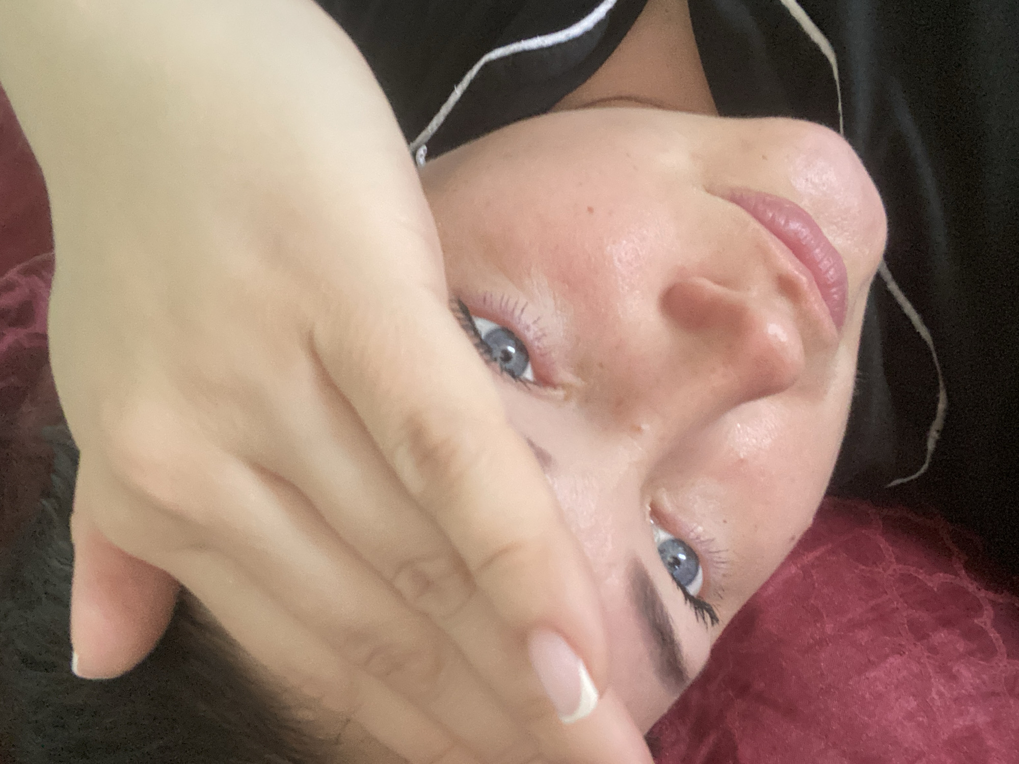 A close-up, angled selfie shows a woman partially obscuring her face with her hand. Her skin appears fair, with blue eyes looking toward the camera. She has manicured nails with a French tip. Her face is positioned above a dark red, textured surface that might be fabric. She is wearing a dark-colored garment with a white trim along the collar or neckline. The background is out of focus and does not provide any information that would allow identification of the location as Sochi or any other place.