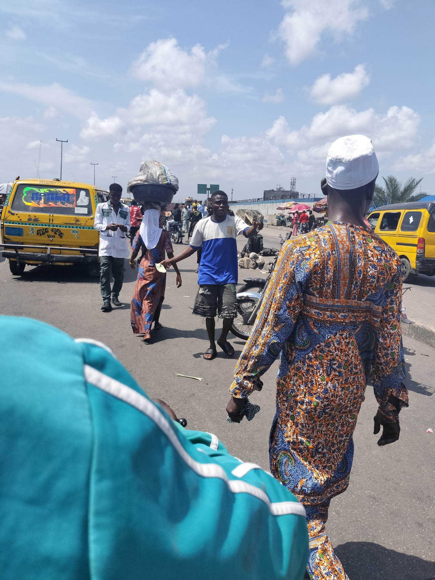 The image captures a bustling street scene in Lagos, Nigeria, under a bright, partly cloudy daytime sky. The perspective is from behind a person wearing a teal/turquoise jacket with white stripes, whose shoulder and back are visible in the foreground.
In the midground, a person wearing a traditional patterned long-sleeved tunic in shades of blue, orange, and white, with a white skullcap (kufi), walks away from the camera. To their left, several other individuals navigate the street. A woman, dressed in a long patterned gown and a white head covering, carries a large basin, wrapped in light material, on her head. She walks past a yellow minibus (commonly known as a 'danfo' in Lagos) adorned with colorful graffiti and stickers on its rear window, including the word "FREEDOM" and "AWE OPE 2000" on the bumper. Another man in a white shirt and dark trousers walks near the bus, while a man in a blue and white t-shirt and shorts (with "Barbie" visible on one leg) gestures with his hand.
In the background, another yellow vehicle is partially visible, along with more pedestrians, market stalls with umbrellas, and a blue wall with structures beyond. The scene conveys the lively and dynamic atmosphere of a typical Nigerian urban street, with people engaged in daily activities, including street vending. The sunny weather and clear sky suggest mid-day.