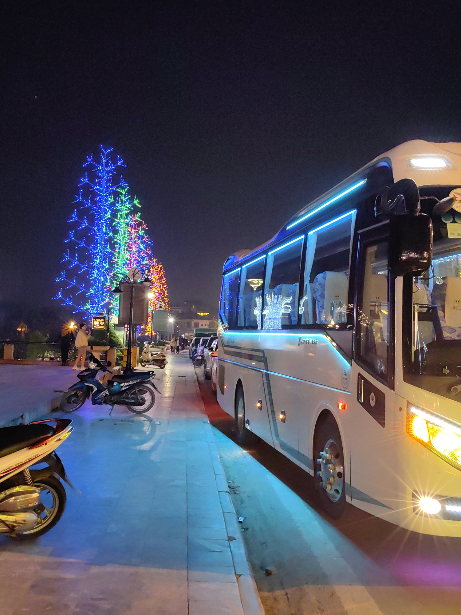 This nighttime image depicts a lively urban scene in Sa Pa, Vietnam. A large, white passenger bus, illuminated by its own bright headlights and prominent blue LED strip lighting along its side windows and roofline, dominates the right foreground, parked parallel to a paved sidewalk. Its interior shows light-colored seats and subtle decorative elements.
On the left, a row of large, abstractly-shaped trees, intricately decorated with vibrant blue, green, and red LED lights, stretches into the distance, providing significant ambient illumination. Several motorcycles are parked on the sidewalk: a dark blue or black motorcycle is in the midground, and the front wheel and partial body of a white and silver motorcycle are in the immediate left foreground.
In the midground, between the parked motorcycles and the illuminated trees, multiple individuals are visible on the sidewalk, some standing and others walking. One person in a light-colored jacket is clearly visible near a streetlamp which features a "WC" sign. Further down the street, additional vehicles, including cars and scooters, are parked. The environment consists of a dark night sky, a well-lit paved sidewalk, and a street curb, with reflections of the artificial lighting visible on the ground, suggesting a wet or reflective surface. Distant buildings and lights are faintly discernible in the background.