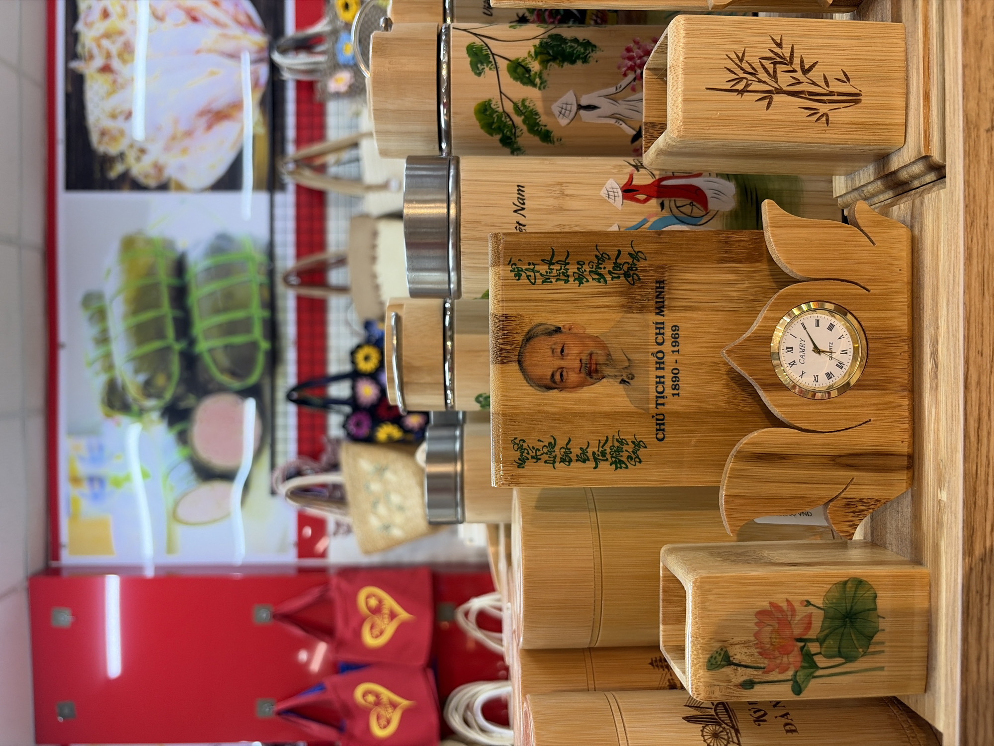 This image depicts a retail display in a store, likely a souvenir shop, in Đà Nẵng, Vietnam. The foreground features an assortment of bamboo-crafted items, including cylindrical containers, a pen holder decorated with a lotus flower, and a unique decorative piece holding a small analog clock and featuring a portrait of Ho Chi Minh, along with Vietnamese script. Behind these items, more bamboo cylinders are visible, some adorned with illustrations of Vietnamese women in conical hats and landscapes. In the background, a red wall with hanging hooks displays two red tote bags with yellow heart designs. Further back, a display wall showcases large photographs of Vietnamese food items, specifically what appear to be traditional rolled cakes. Several woven bags with floral embellishments are also present on shelves. The lighting suggests an indoor setting with overhead illumination. There are no people visible in the image.