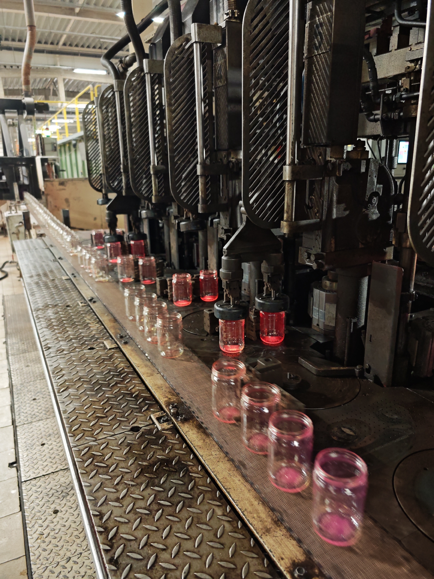 The image depicts a glass manufacturing line, likely in an industrial setting. A long row of clear glass jars with pink rims and bottoms move along a conveyor belt. Above the jars, a series of metal machinery units are positioned to perform some operation on the glass. The jars appear to be hot, glowing red internally at some points along the line. The conveyor belt is made of a textured metal surface. The location in the city of Pleven, Bulgaria is unknown.