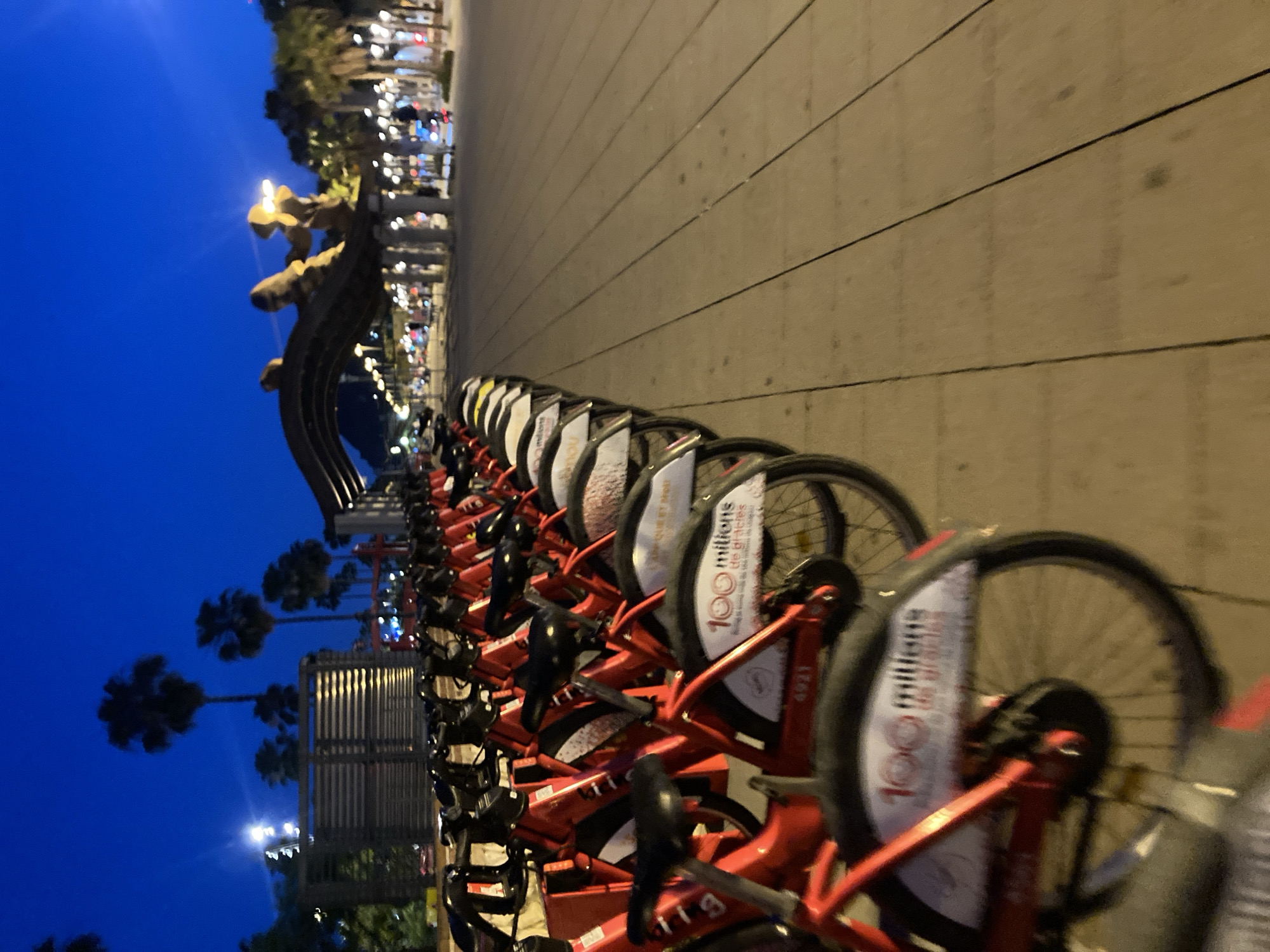 The image depicts a row of red bicycles parked along a wooden promenade at dusk. The bicycles, likely part of a city bike-sharing system, are neatly lined up, their frames and handlebars reflecting the ambient light. Some of the bicycles feature signage on their fenders, one of which reads "100 millions de gracies." In the background, palm trees and architectural elements suggest a Mediterranean setting, specifically the Barceloneta boardwalk in Barcelona, Spain, known for its public spaces and bike lanes. The sky is a deep blue, transitioning from twilight to night, with streetlights and lights from buildings visible in the distance. The overall impression is one of a quiet, urban evening, with the bicycles awaiting their next riders.