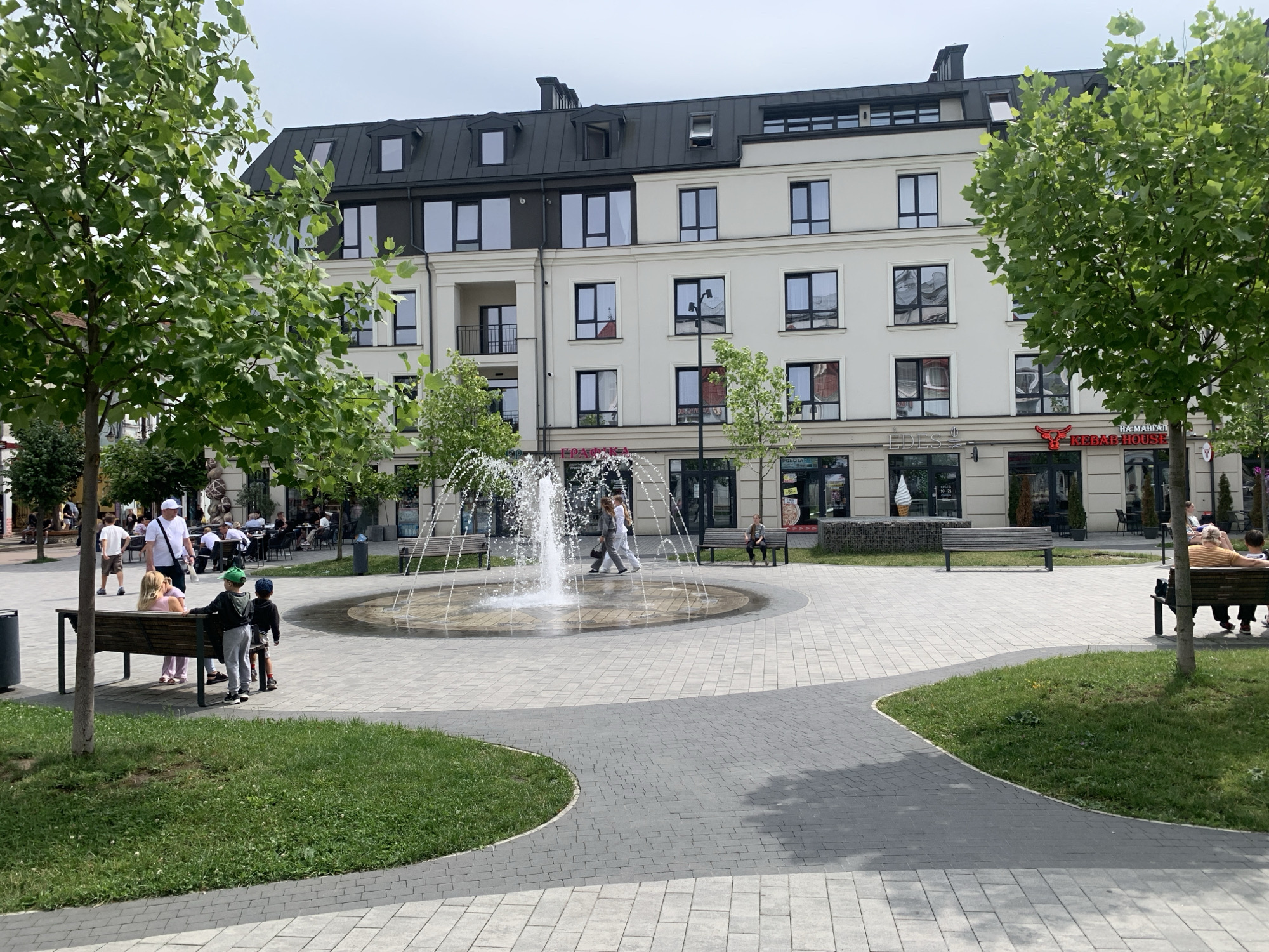 In the city of Kolomyia, Ukraine, a modern building with a dark pitched roof and white facade stands prominently, featuring several windows and dormer windows. In front of it, a public square is paved with light-colored bricks and features a circular fountain with water spraying upwards. Green grass is interspersed with the paving, and trees with lush green foliage are visible. Several benches are placed around the fountain and in the grassy areas.
On the left side of the image, two young children, one wearing a green baseball cap, are standing near a bench, facing away from the camera. A woman is seated on the bench, facing the children. To the left of them, a man in a white t-shirt and shorts, and another person in a white t-shirt and shorts, are walking. Further back, several people are seated at tables outside a building.
On the right side of the image, two people are seated on a bench, facing away from the camera. Another person is seated on a bench closer to the fountain, facing towards the fountain. The building in the background has several storefronts, including one with a "Kebab House" sign. The sky is overcast. The overall scene depicts a pleasant, modern urban public space.