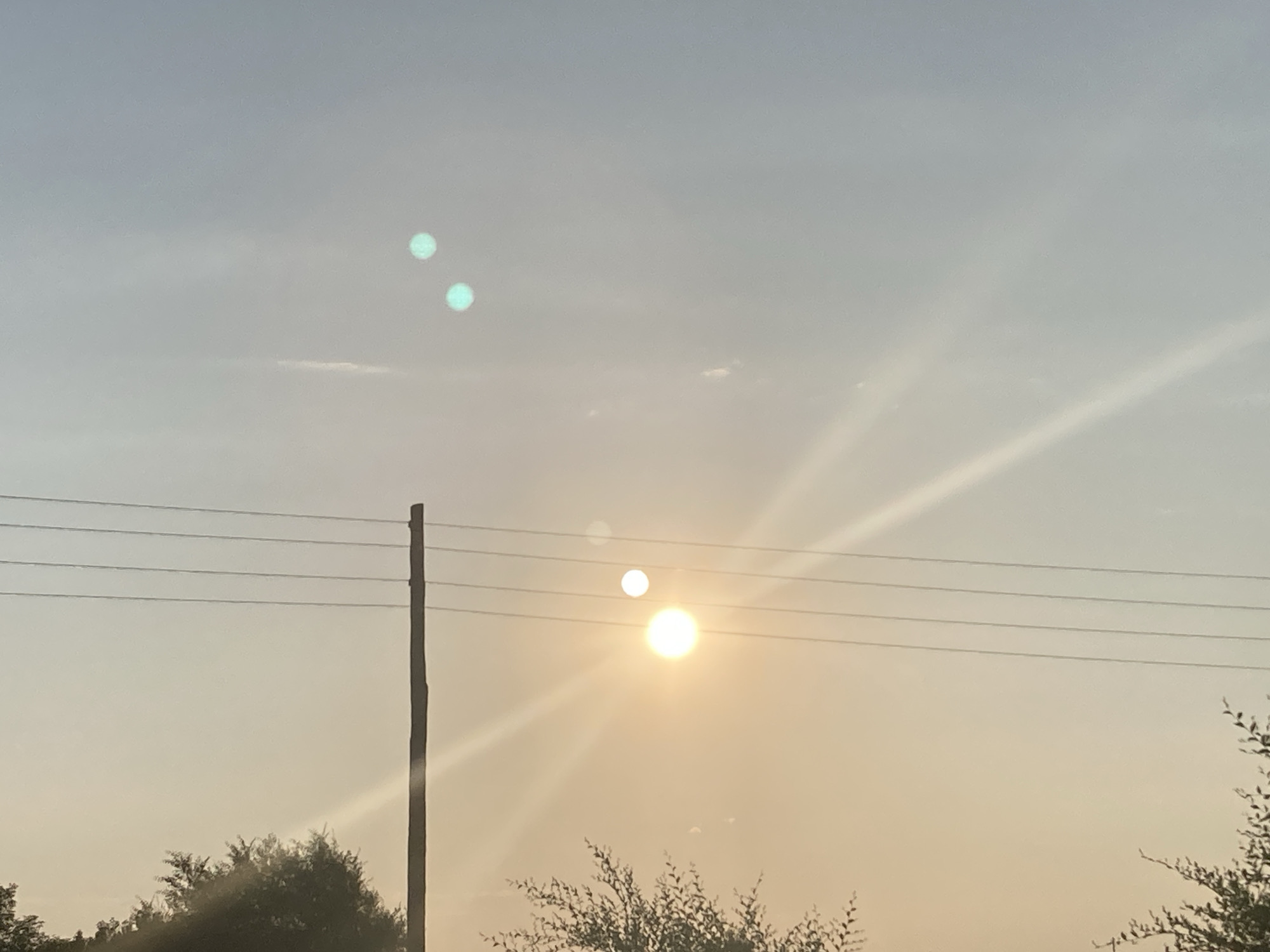 A power pole stands to the left of the frame, with four parallel wires stretching across it. The sun is low in the sky, casting a warm glow and creating lens flares. Two of these flares appear as bright, circular orbs, with one significantly larger and more intense than the other. There are also two smaller, teal-colored orbs higher in the sky, likely due to lens artifacting. The background consists of a hazy sky with faint clouds. The bottom of the frame shows the dark silhouettes of trees and foliage. The scene is set during the day, under an overcast or hazy sky, with the sun being a prominent element. The location is specified as the city of Rakity, Russia.