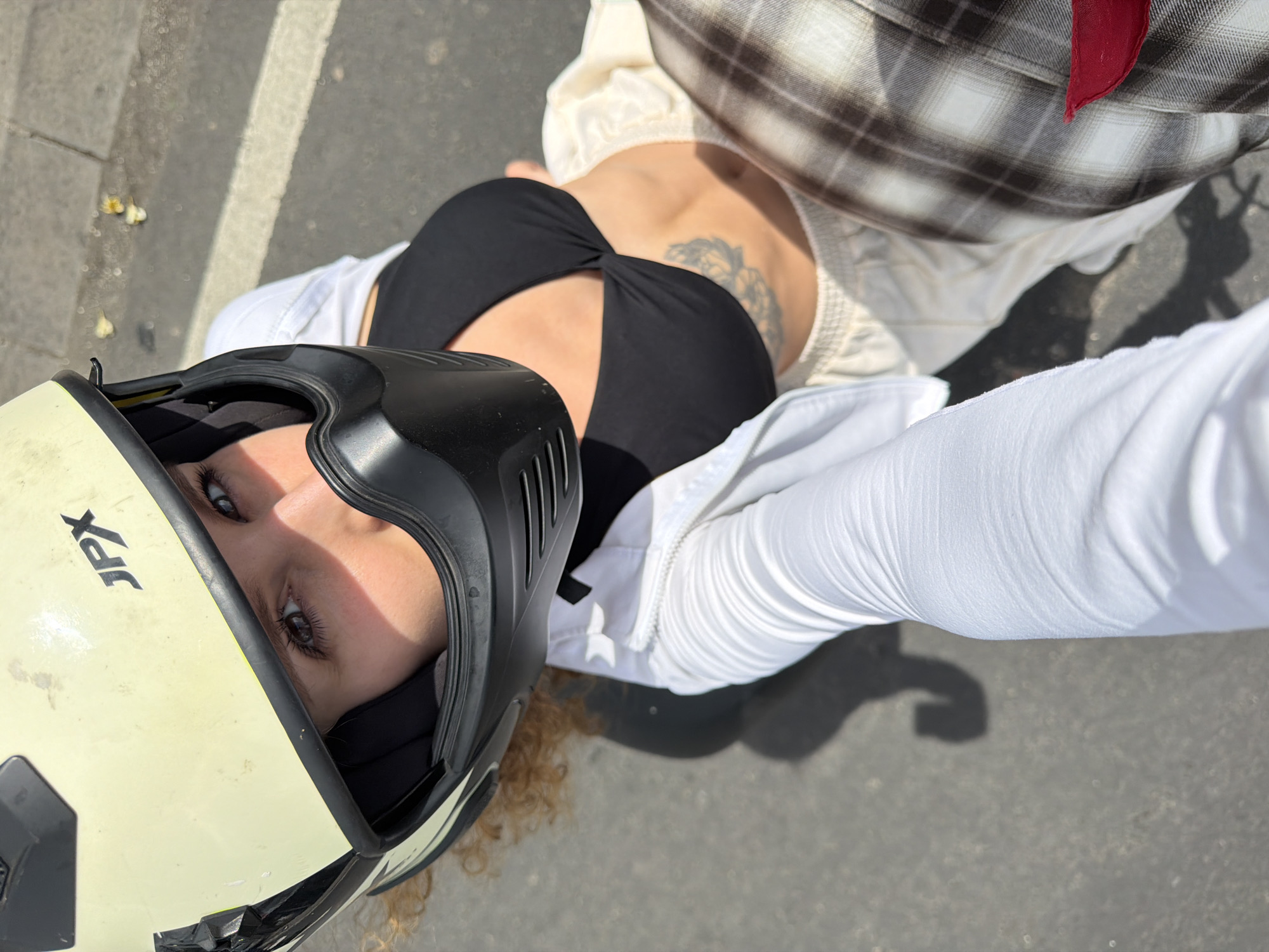 A woman wearing a yellow and black helmet and a black bikini top is looking up at the camera. She has blonde curly hair and a tattoo on her chest. She is wearing a white open jacket and light-colored pants. The background appears to be a street in North Kuta, Indonesia, with asphalt and a white line painted on the ground. The scene is illuminated by bright sunlight, casting shadows on the ground.