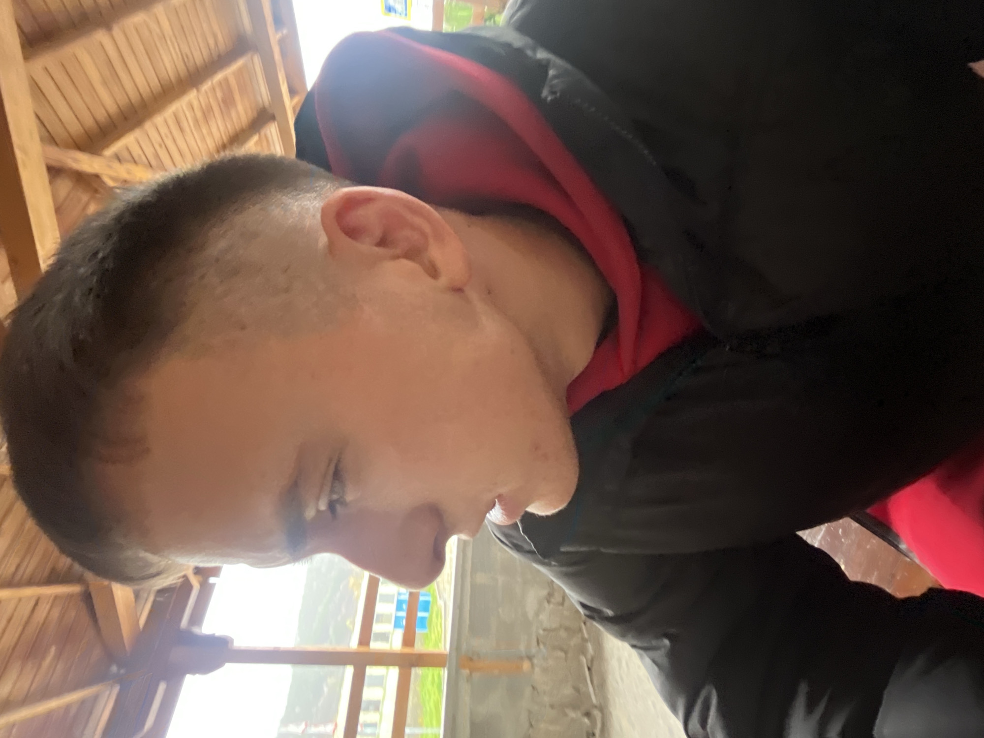 A man with short, dark hair is seen from the side, facing left. He's wearing a red hoodie under a black jacket. The background includes a wooden structure, possibly a roof, and an exterior view with green vegetation and buildings. The image was taken in the city of Asenovgrad, Bulgaria.