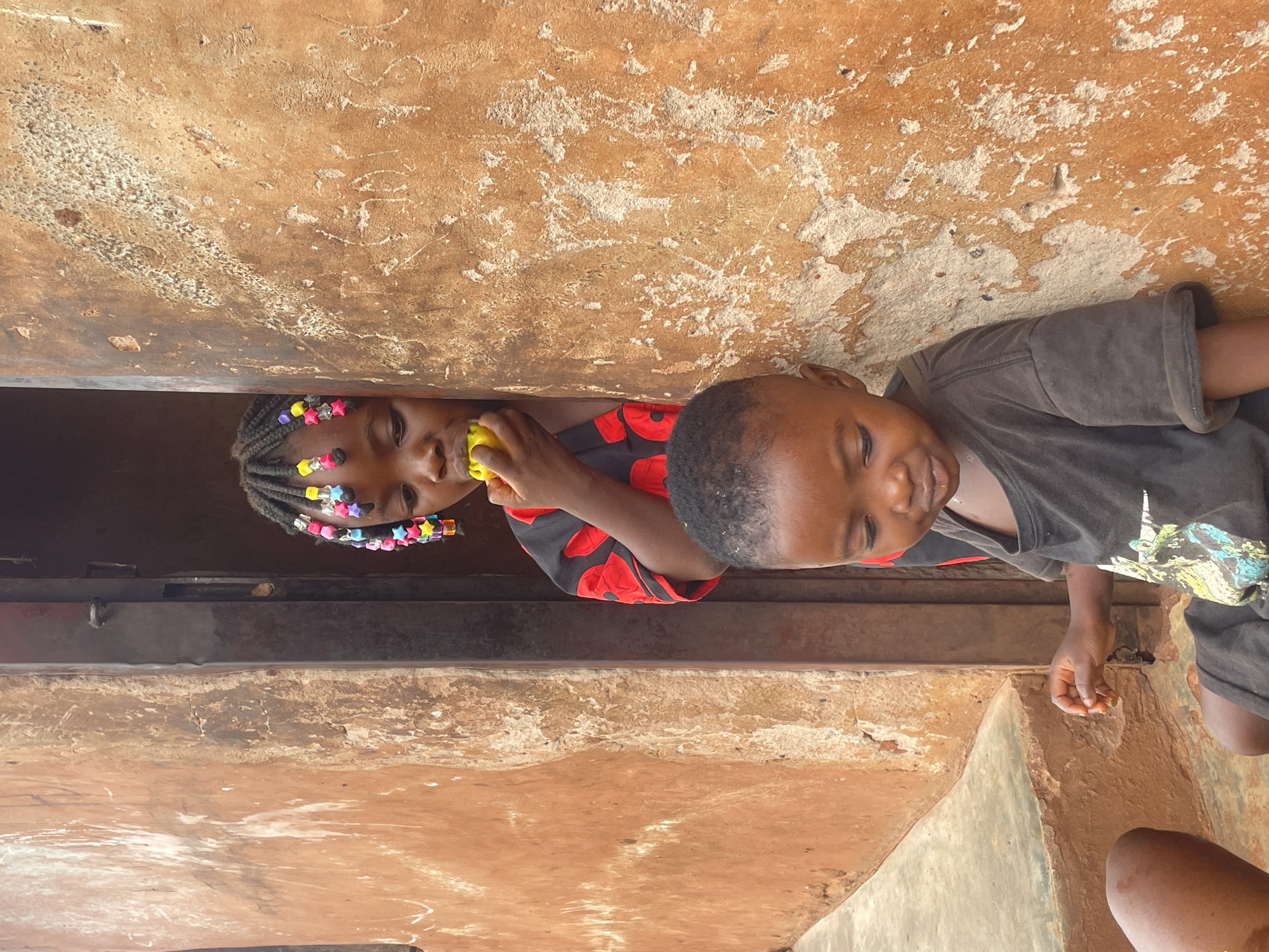 Two children, a girl and a boy, are positioned against a wall in what appears to be Ogbe, Nigeria. The girl, on the left, has dark skin, braided hair adorned with colorful beads, and is holding a yellow object in her hand. She is wearing a red and black patterned shirt. The boy, on the right, has short hair and a dark-colored t-shirt. He is smiling. The wall behind them is textured and painted with orange and white hues. A dark metal bar is positioned between them and the wall. The setting suggests an outdoor environment, possibly a doorway or entrance to a building.