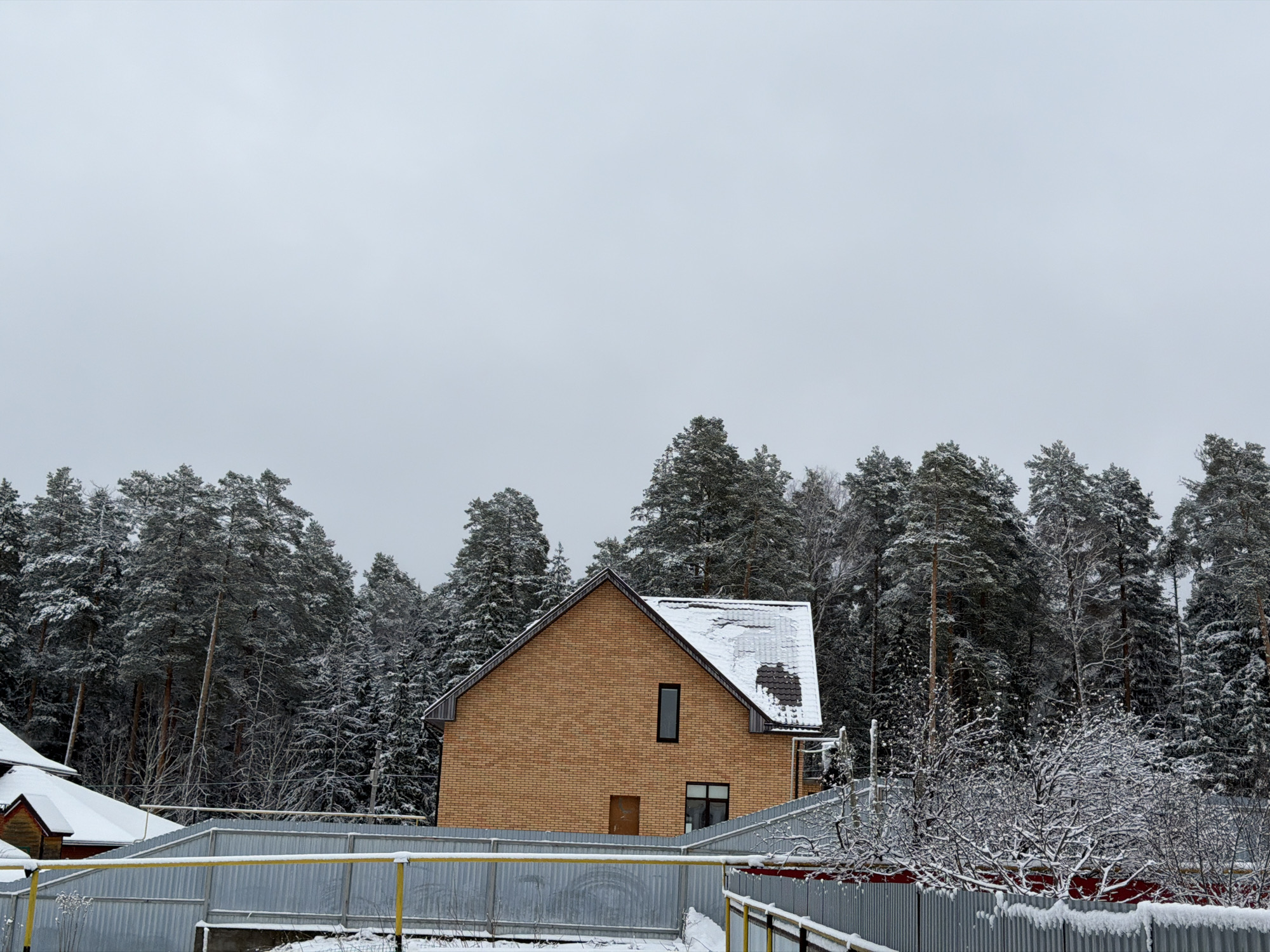 The image depicts a two-story brick house with a dark roof covered in patches of snow, situated in a snowy environment possibly during winter. The house has a small rectangular window on the second floor and a door with a partially visible window on the first floor. The house is surrounded by tall, snow-covered evergreen trees, possibly a forest. In the foreground, there's a corrugated metal fence partially covered in snow. The sky is overcast, contributing to the somber atmosphere. There are also parts of other houses in the vicinity. The location is Chaikovsky, Russia. There are no people visible in the scene.