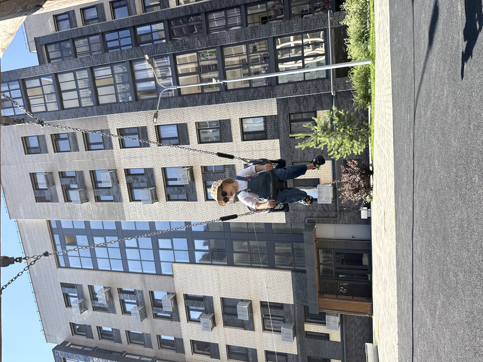 In the city of Rusanovo, Russia, a young child wearing denim overalls, a white t-shirt, a straw hat, and sunglasses sits on a swing. The child is positioned in the foreground, with the swing's chains extending upwards and out of frame. Behind the child, a modern residential building with a façade of light-colored and dark-colored brick, and numerous windows, dominates the scene. The building appears to be a multi-story apartment complex. To the right of the building, a young pine tree and other green bushes are visible. The ground in the foreground consists of dark asphalt. The overall impression is that of a contemporary urban playground setting.