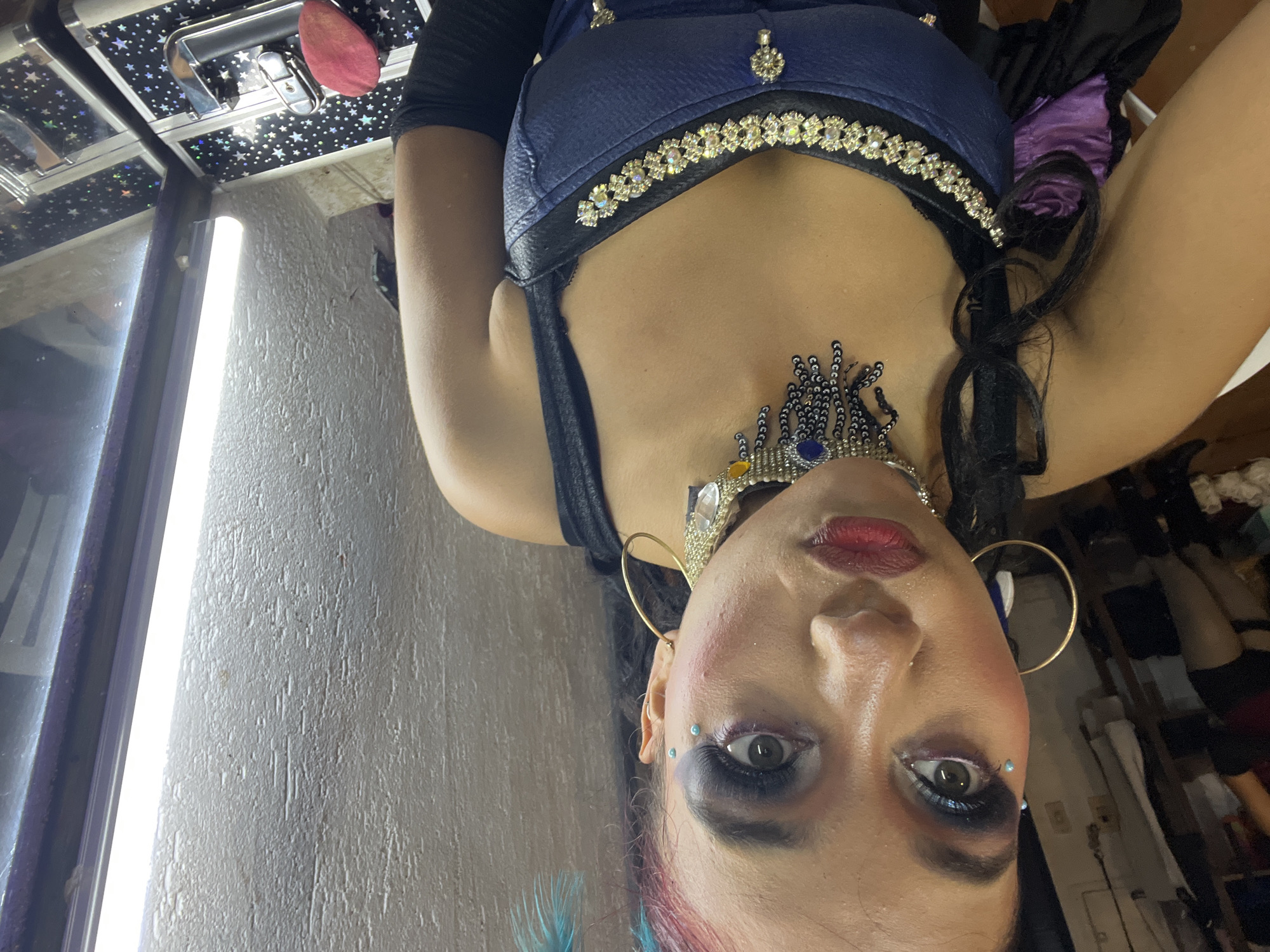 The image shows a close-up, head-and-shoulders selfie of a woman with elaborate makeup and costume. She has dark hair with pink and blue streaks, dramatic eye makeup including blue eyeshadow and false lashes, and blue rhinestones on her cheekbones. She is wearing a jeweled choker and a blue and black costume with rhinestone embellishments. In the background, to the left, are clothing racks with items of clothing. To the right, a white wall with textured details is visible, as well as a bright vertical light fixture and a reflection in a mirror of what appears to be a makeup case. The overall impression is of a performer or someone in costume, possibly backstage. The location is unknown.