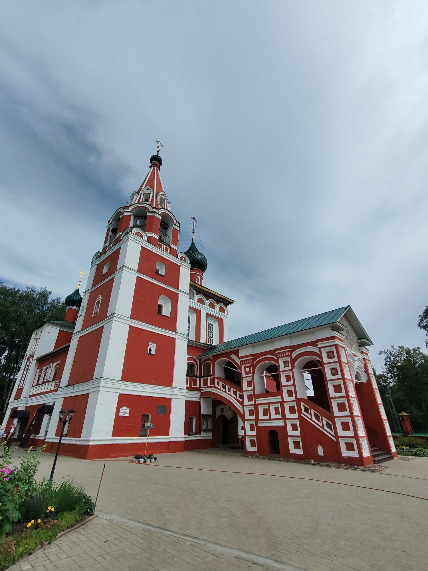 The image shows a wide-angle, low-angle view of a red and white Orthodox church in Rusanovo, Russia, under a cloudy sky. The church features a tall bell tower with a red spire topped by a golden cross. Adjacent to the tower is a section of the church with green domes and crosses, and a red entrance with white decorative arches and a green roof. The church has intricate white trim against the red facade. In the foreground, a paved walkway leads to the church, bordered by a flowerbed with pink and yellow flowers. There are two lampposts near the entrance and a sign that reads "ВХОД" (Entrance). A few people are visible in the distance near the church entrance. Trees line the background of the scene.