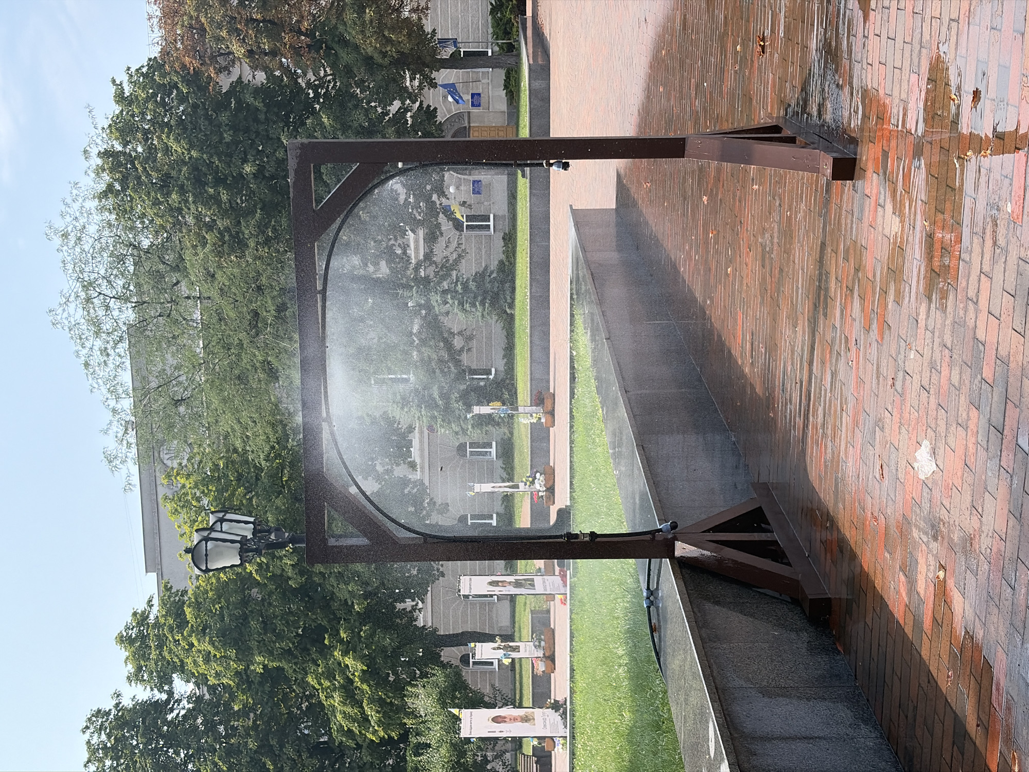 The image shows a metal archway with a misting system in operation, spraying water into the air. The archway is made of dark brown metal and has a triangular support structure at the base of each upright. The mist is creating a translucent veil in front of the archway and on the ground. The ground is paved with reddish-brown bricks that are wet and reflective, with puddles of water in places. To the left of the archway is a low, dark grey stone wall, behind which is a patch of green grass. In the background, there are several tall, thin banners displayed on poles, some with portraits and Ukrainian flags. Beyond the banners, there are trees with lush green foliage and a building with a light-colored facade and windows. A lamppost with two lamps is visible behind the archway, partially obscured by the mist. The sky is clear and blue. The overall scene suggests an outdoor public space, possibly a park or plaza, where the misting system is being used to cool the area.