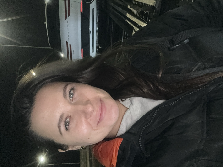A woman with long dark hair and light eyes smiles toward the camera. She wears a gray shirt and a black jacket with an orange collar. Behind her, a white Genesis GV70 car is parked, illuminated by a streetlight. The background is dark, suggesting it is night. There is what appears to be some wooden planks behind her as well.