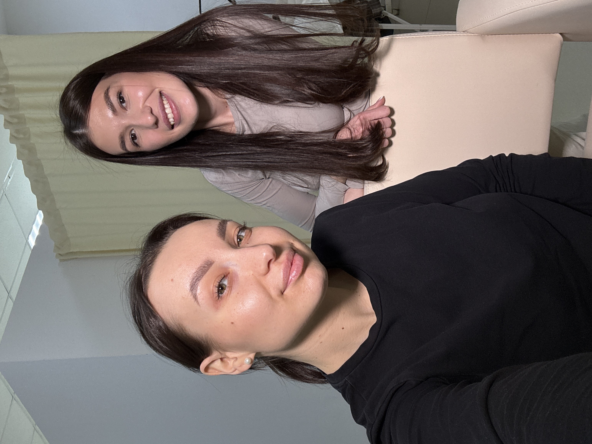 The image shows two women posing for a selfie. One woman is wearing a black long-sleeved shirt and is positioned at the bottom of the frame. The other woman is wearing a light-colored top and has long, dark hair. They are indoors, sitting on a light beige chair. The background includes a light-colored curtain and what appears to be a medical or beauty treatment chair. The location is Chaikovsky, Russia.
