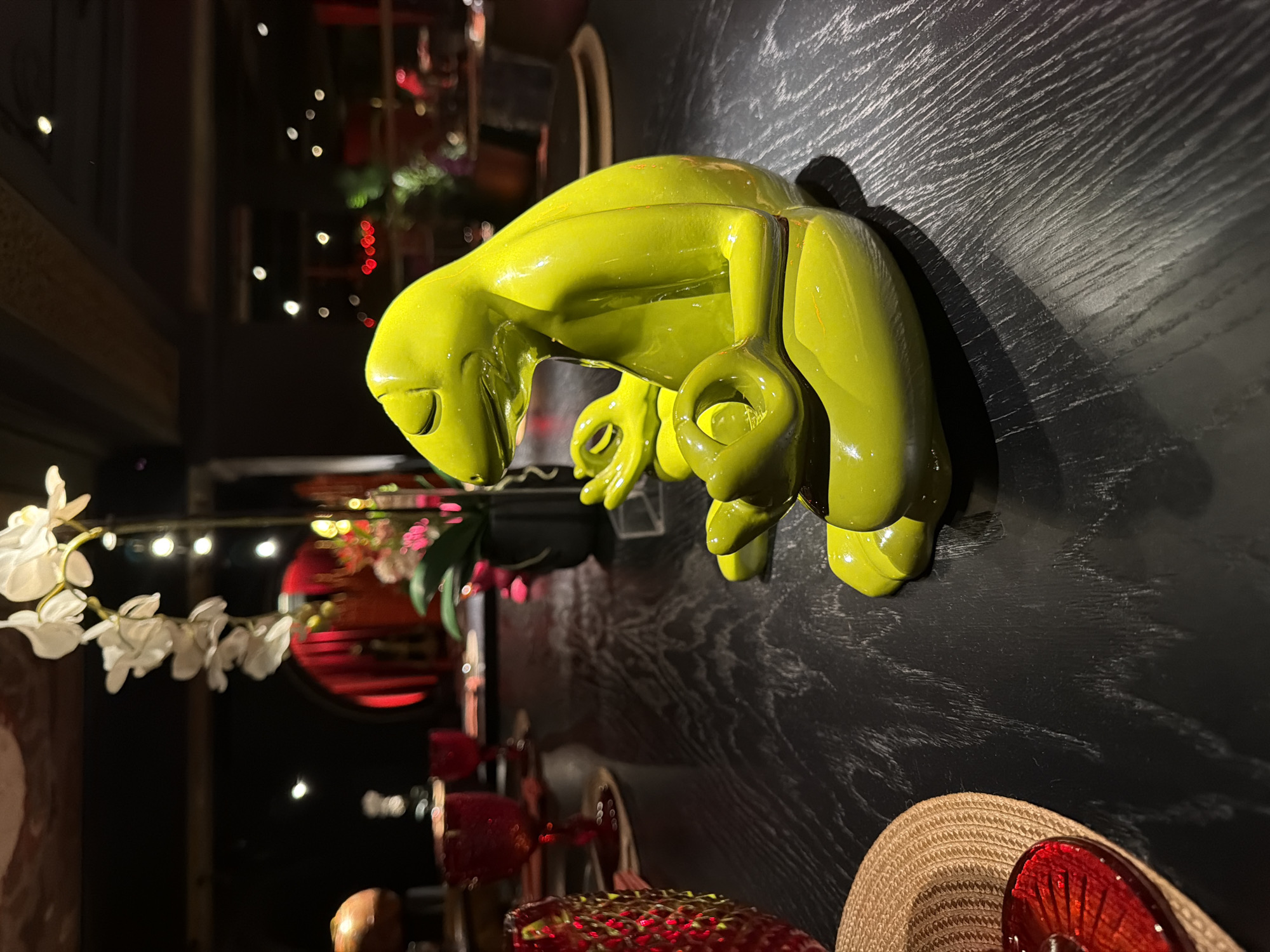 A glazed, chartreuse-colored sculpture resembling an abstract frog hangs on a dark wood paneled wall with a distressed texture. The sculpture is mounted near a shelf or table displaying decorative objects including a light colored straw hat with a red accent, decorative red glass, an artificial white orchid, and wine glasses. The background is out of focus, showing indistinct elements of a room with soft, bokeh-like lights, and what appears to be a red circular structure with floral embellishments. The location might be the Moscow, Russia, but no features clearly indicate the city.